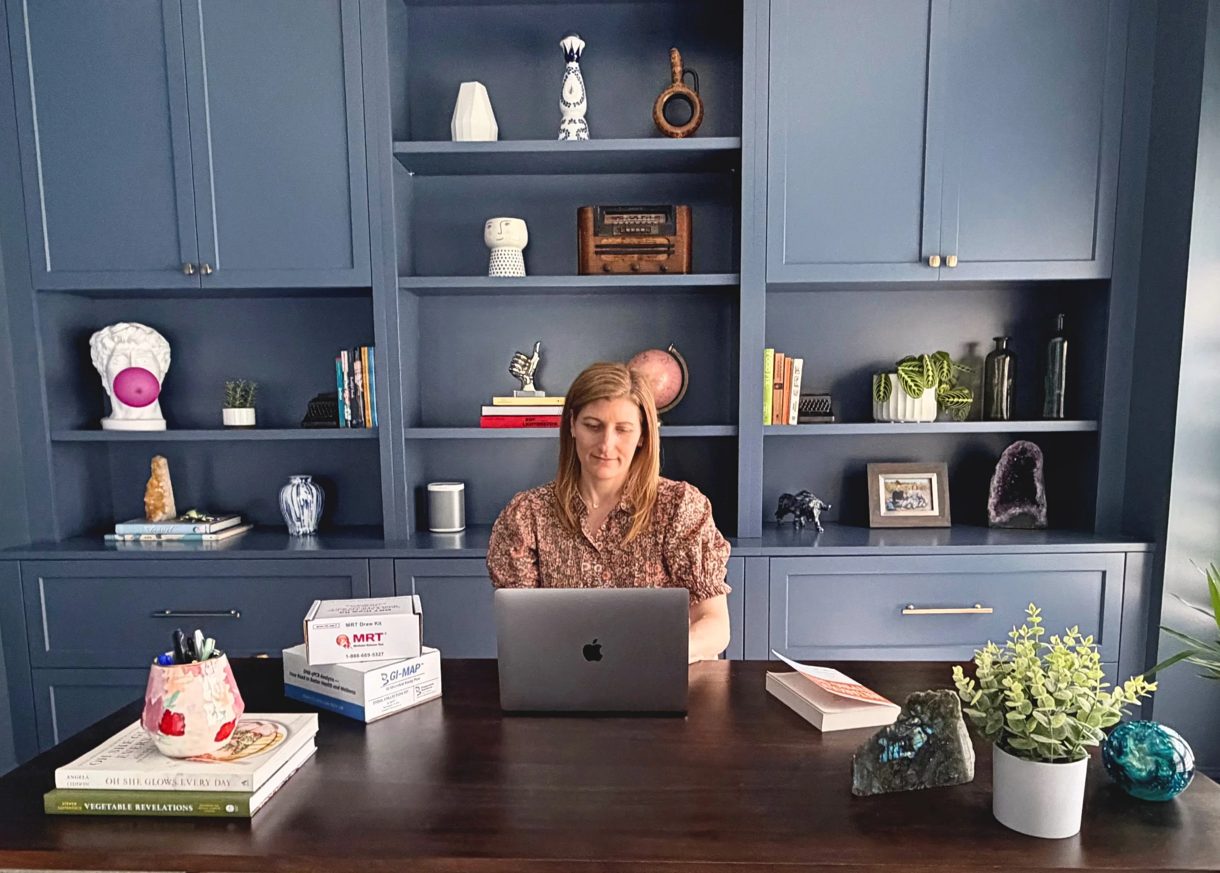 Woman sitting at a desk with a laptop, surrounded by plants, decorative items, books, and office supplies, in front of a blue built-in bookshelf with various decorative objects.