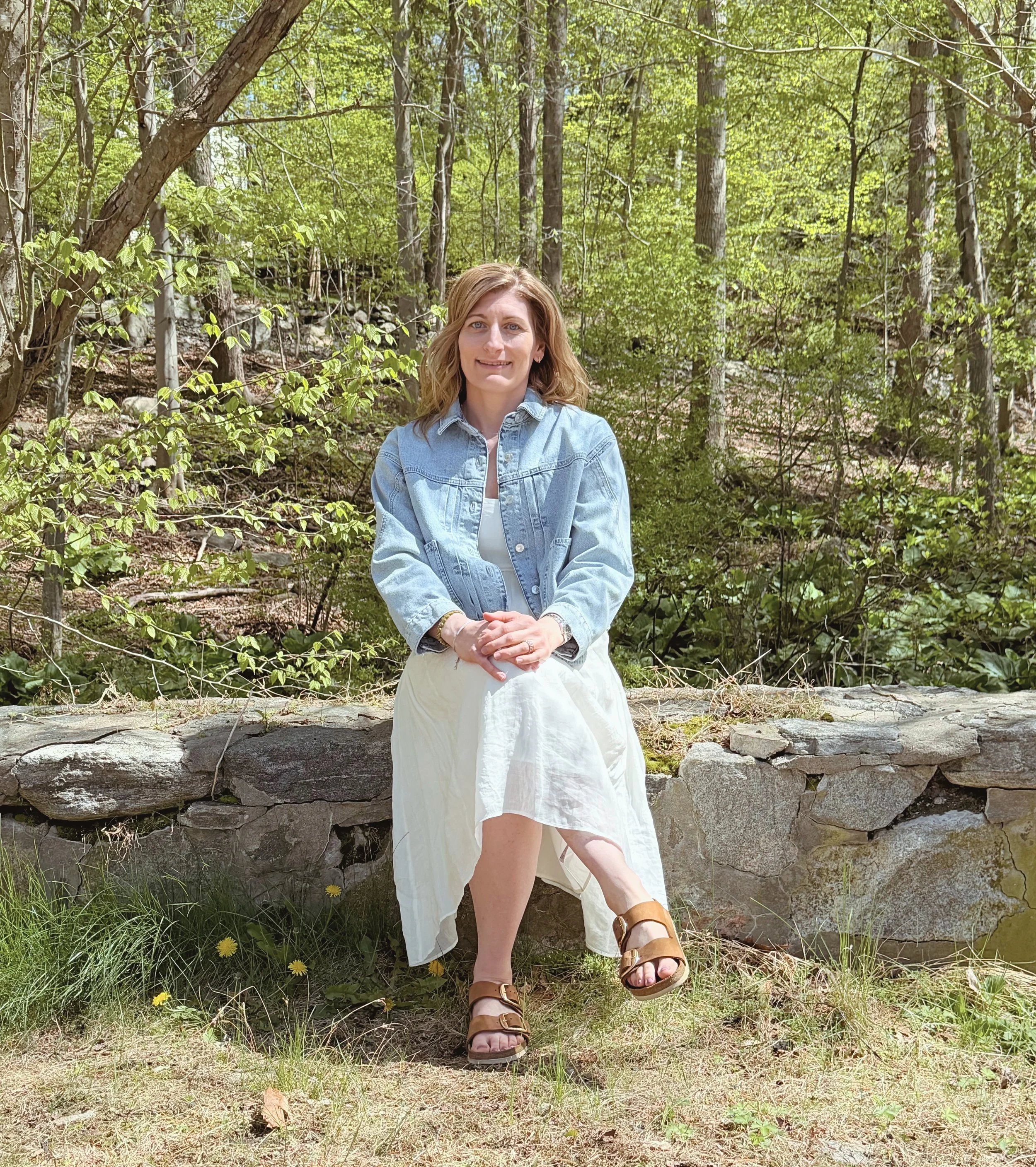 A woman with light brown hair sitting outdoors on a stone ledge in a wooded area, wearing a denim jacket, white dress, and brown sandals, with a natural background of trees and green foliage.