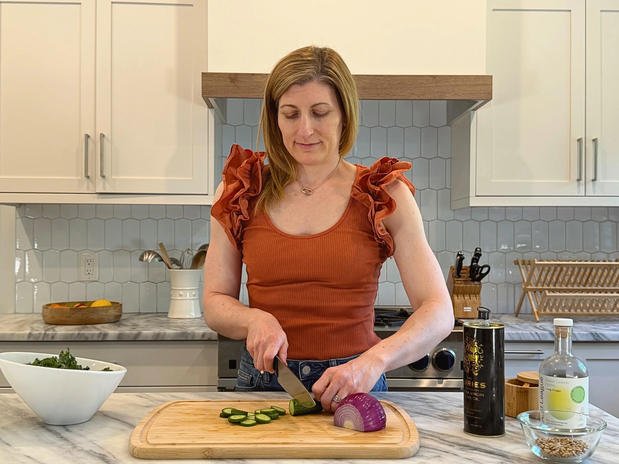 A woman is slicing a cucumber and red onion on a wooden cutting board in a modern kitchen with white cabinets and a hexagon tile backsplash.