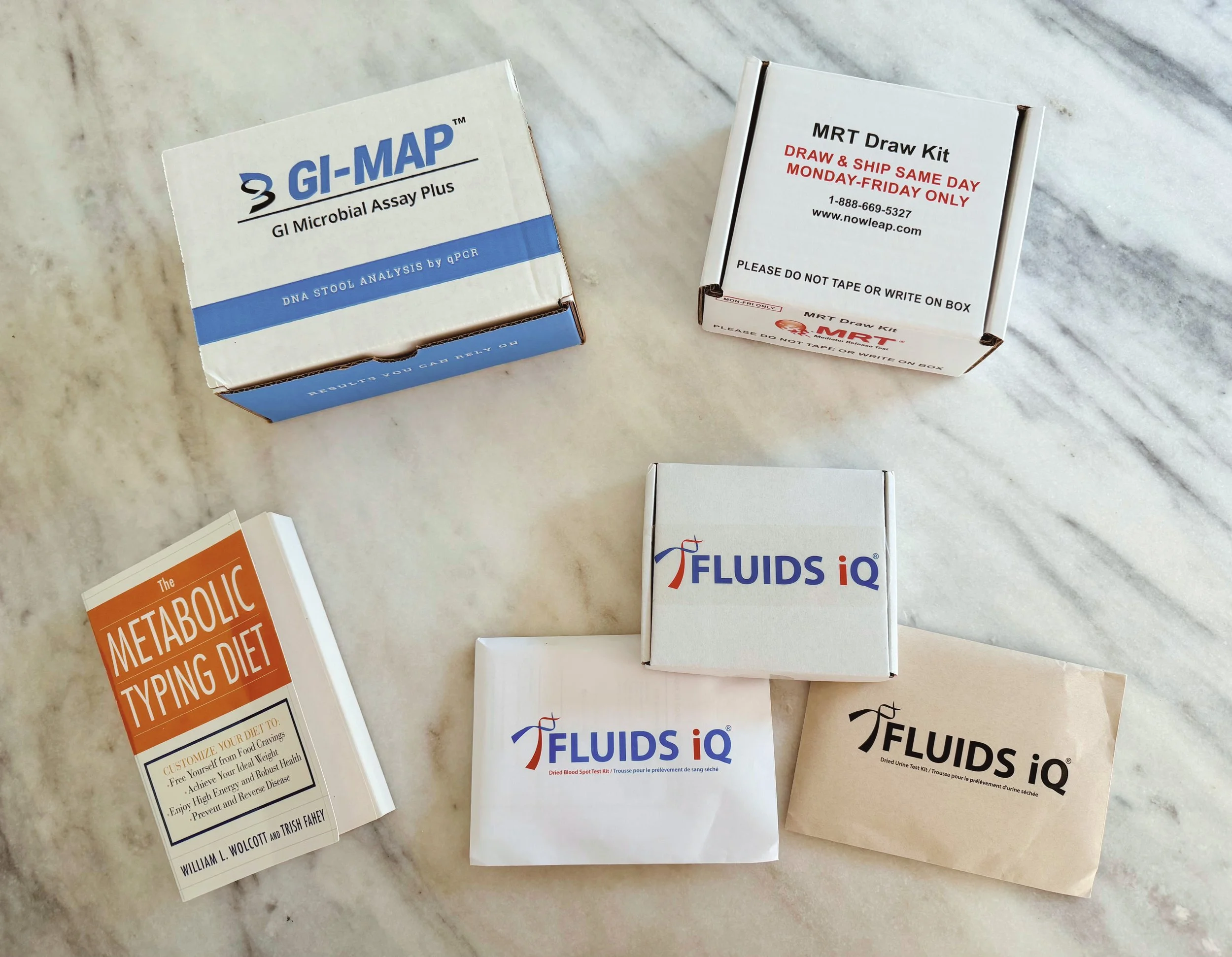 Various health and diet-related informational booklets, including GI Microbial Assay Plus, MRT Draw Kit, and Fluids IQ, spread out on a marble surface.