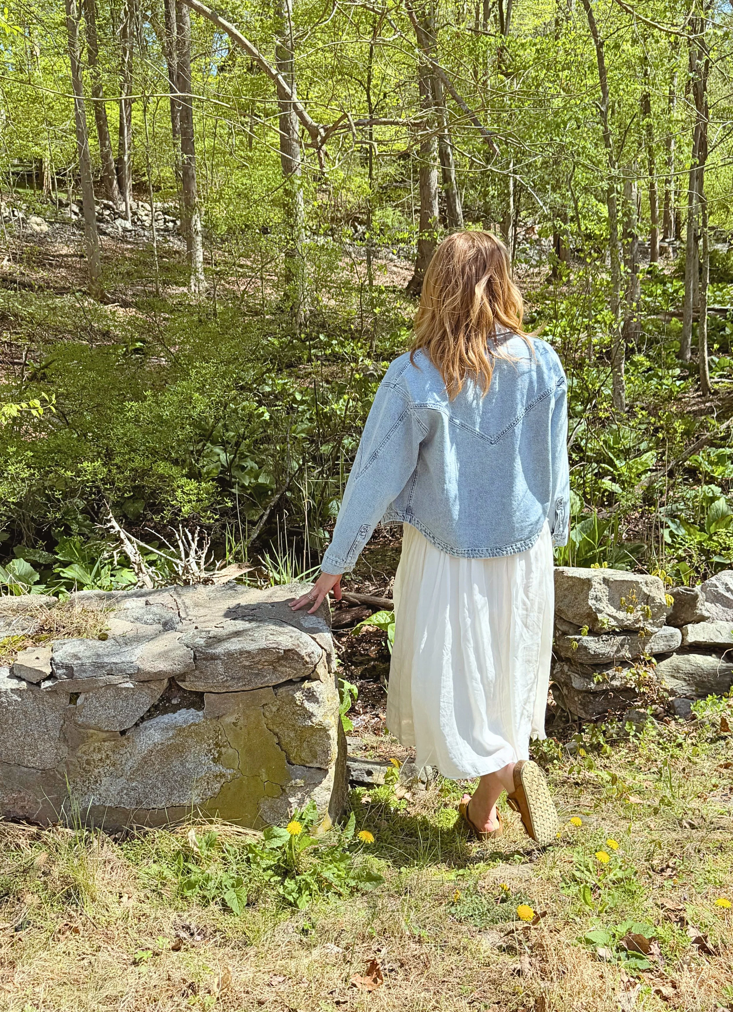 A person with blonde hair wearing a light blue denim jacket and a long white skirt is standing on a rock wall in a green forest, facing away from the camera, with one hand resting on the stone wall and the other down by her side.