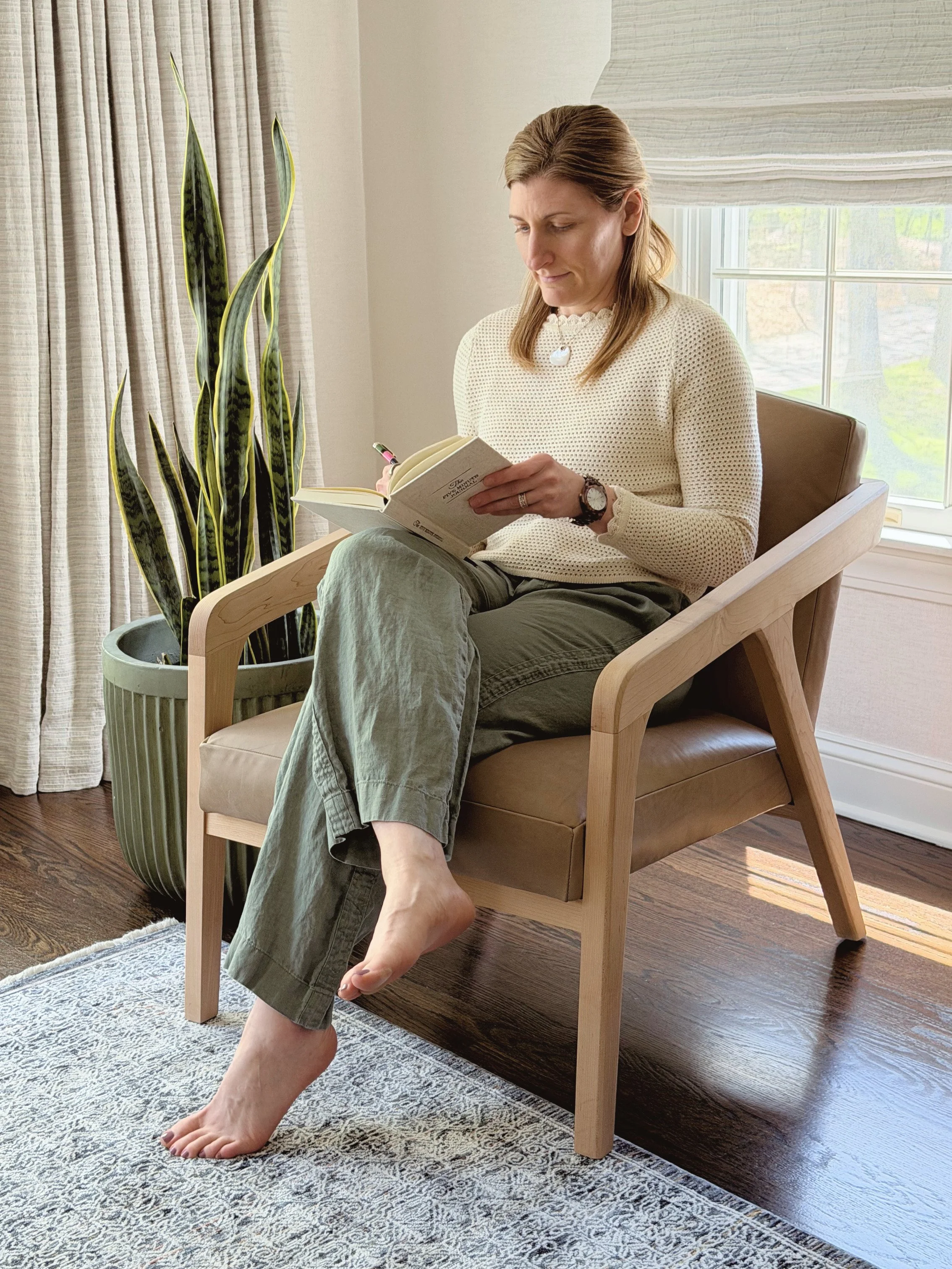 A woman sitting in a wooden armchair near a window, holding a book, with a large potted snake plant beside her and light-colored curtains in the background.