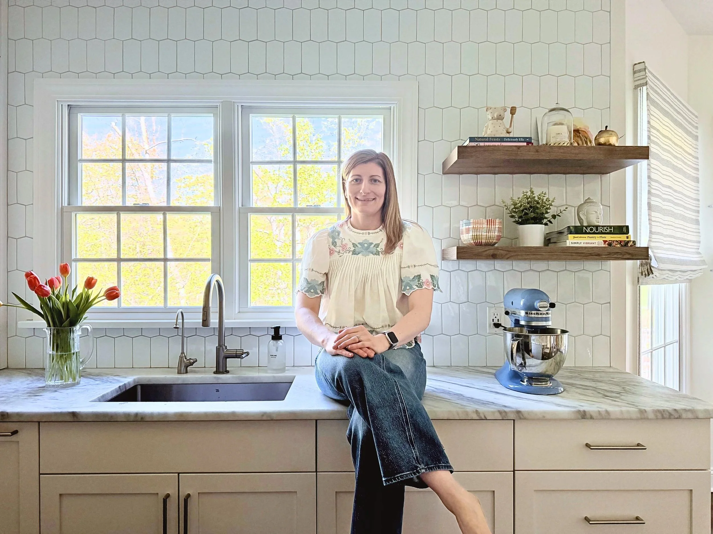 Woman in a cream-colored floral blouse and jeans sitting on a kitchen countertop with her arms crossed, smiling, in a bright kitchen with a window behind her, a vase of tulips, a wooden floating shelf with books, and a stand mixer.