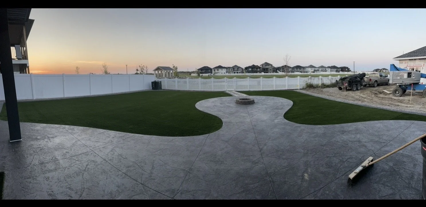 Backyard patio with a circular concrete fire pit, green grass patches, white fence, and a clear sunset sky. Construction vehicles and equipment visible on the right side.