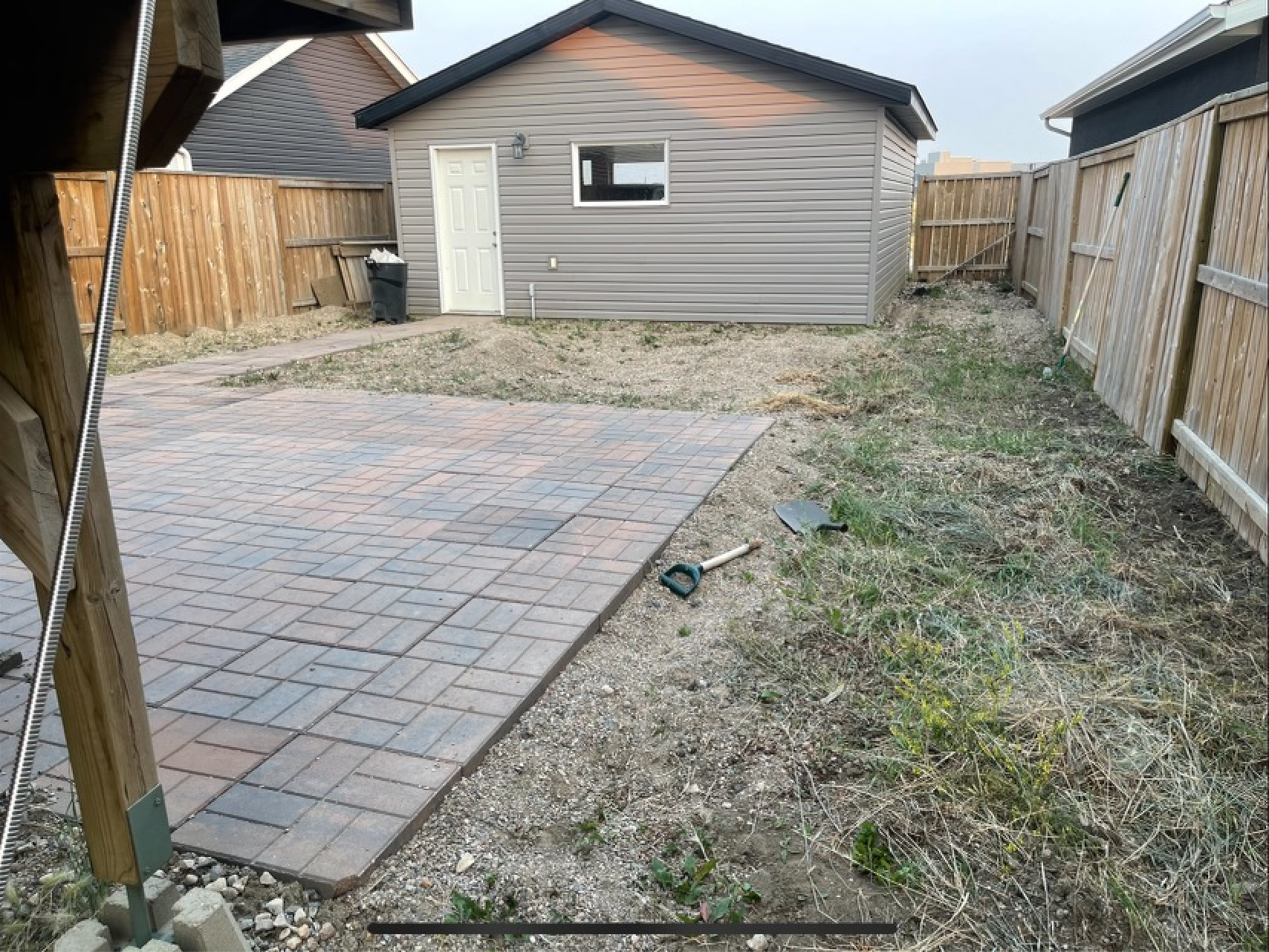 Backyard with newly paved patio, wooden fence, small shed, and gardening tools
