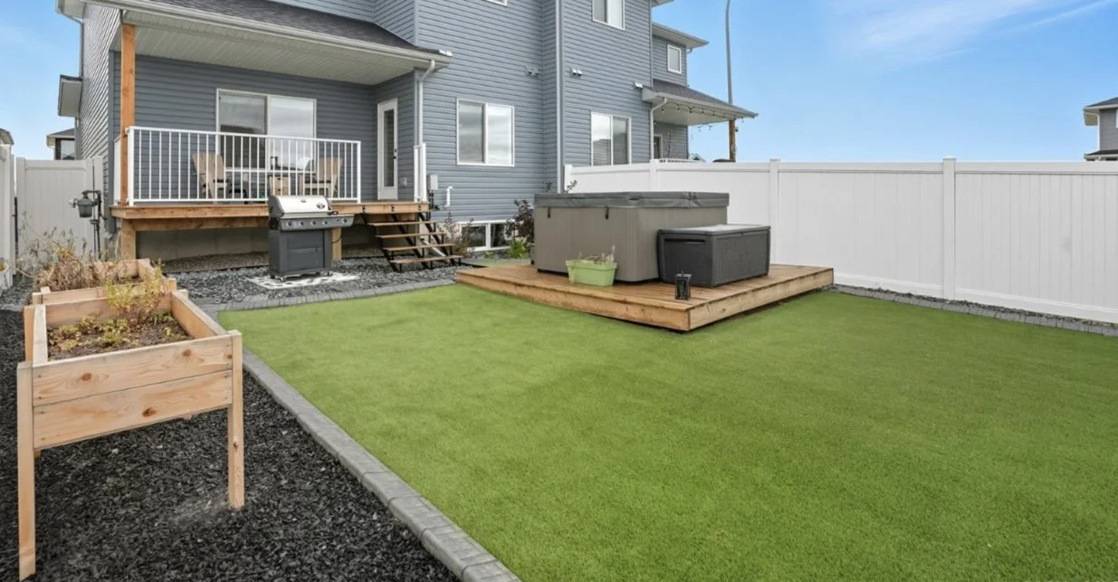 Backyard yard with a lawn, wooden deck with hot tub, grill, and outdoor furniture, raised garden bed, and white privacy fence, blue house in background.