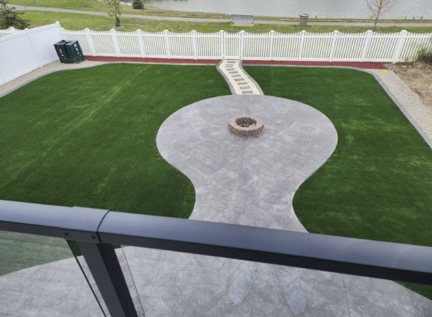 An outdoor backyard with a grass lawn, a paved circular patio with a fire pit in the center, a gravel pathway leading to a white fence, with trash bins in the corner and a body of water in the background.
