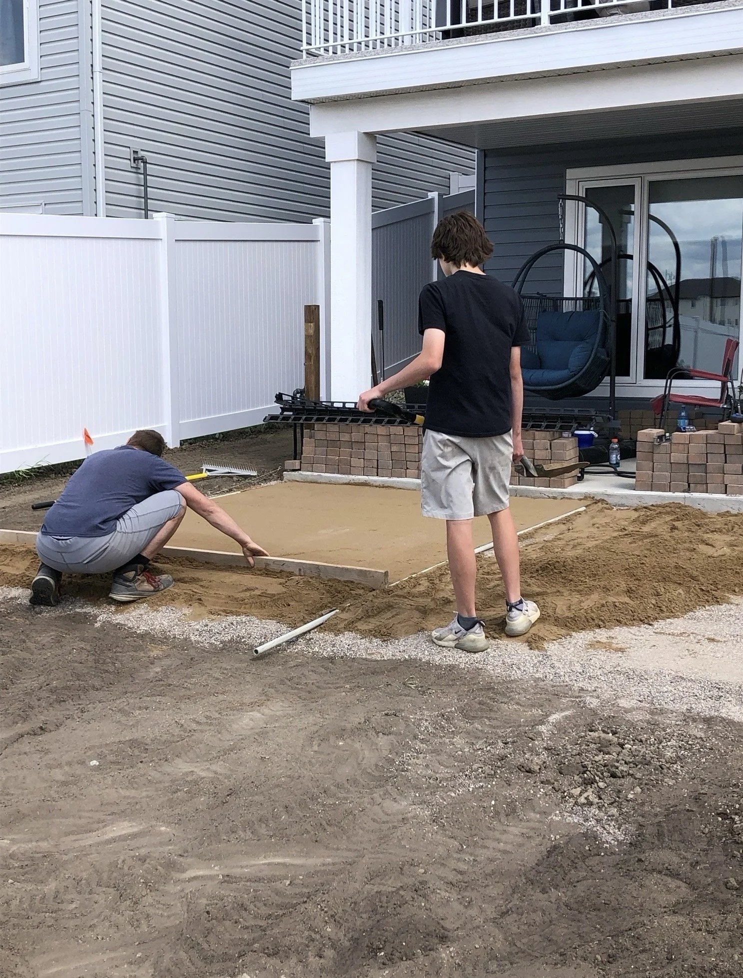 Two young men working on a backyard landscaping project, spreading and leveling soil. One is kneeling and the other standing, with a house, sliding glass door, swing chair, and patio furniture in the background.