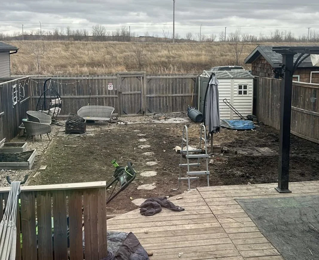 A backyard in the process of being landscaped with a dirt area, stepping stones, a small wooden deck, outdoor furniture, a plastic storage shed, and a backyard with a hill and utility poles in the background.