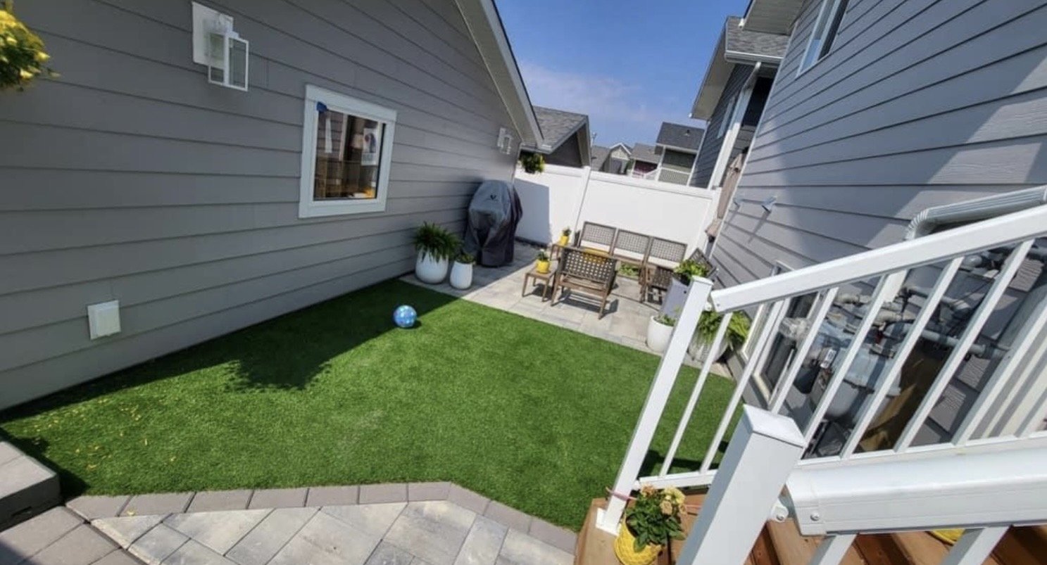 Backyard patio with artificial grass, outdoor furniture, potted plants, and a barbecue grill enclosed by white fence and neighboring houses.