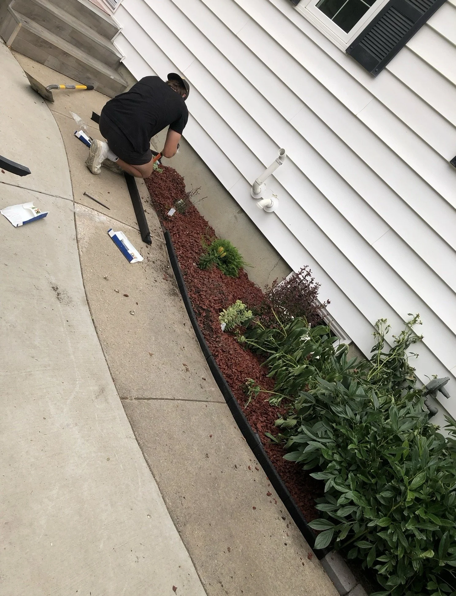 A person kneeling down, working on a garden bed beside a house, using tools and surrounded by gardening supplies.