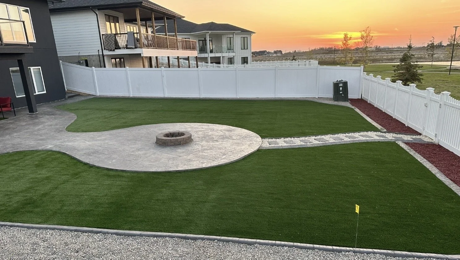 Backyard with a fire pit, artificial grass, a concrete patio, a white fence, and sunset in the background.