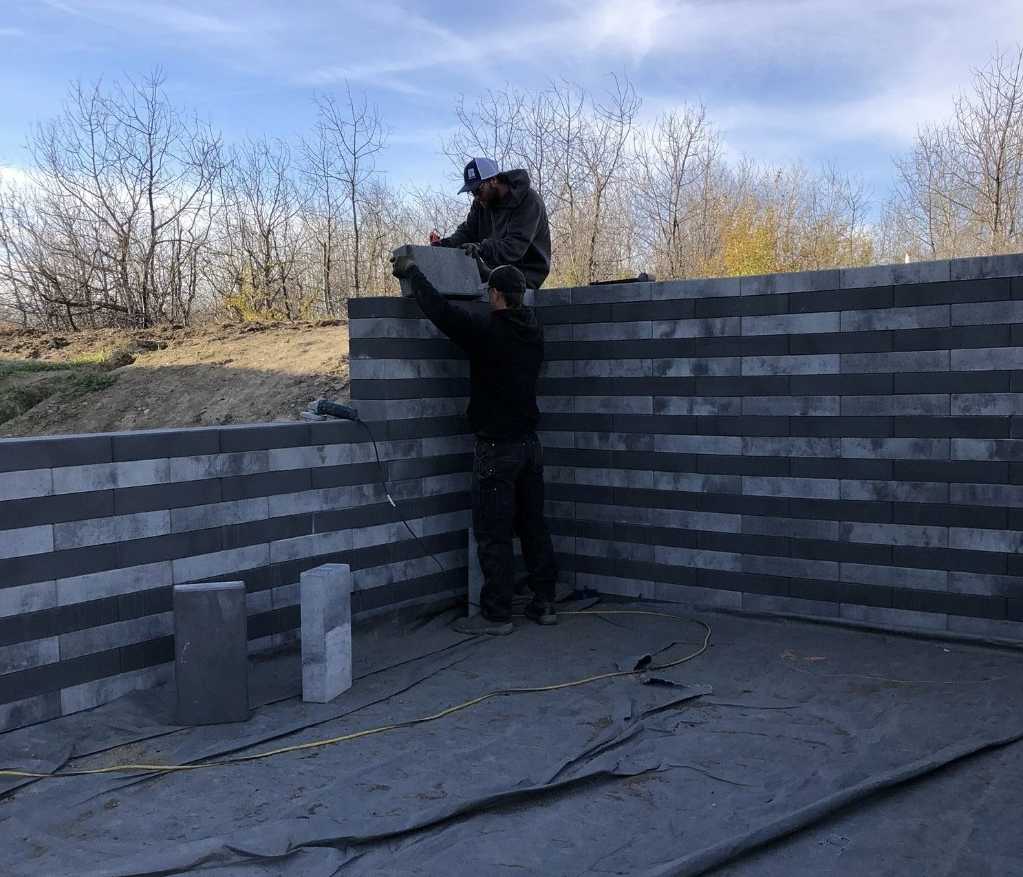 Two workers constructing a gray brick wall outdoors during daytime, with one standing and passing a brick to the other on top of the wall, while trees with bare branches and a blue sky are in the background.