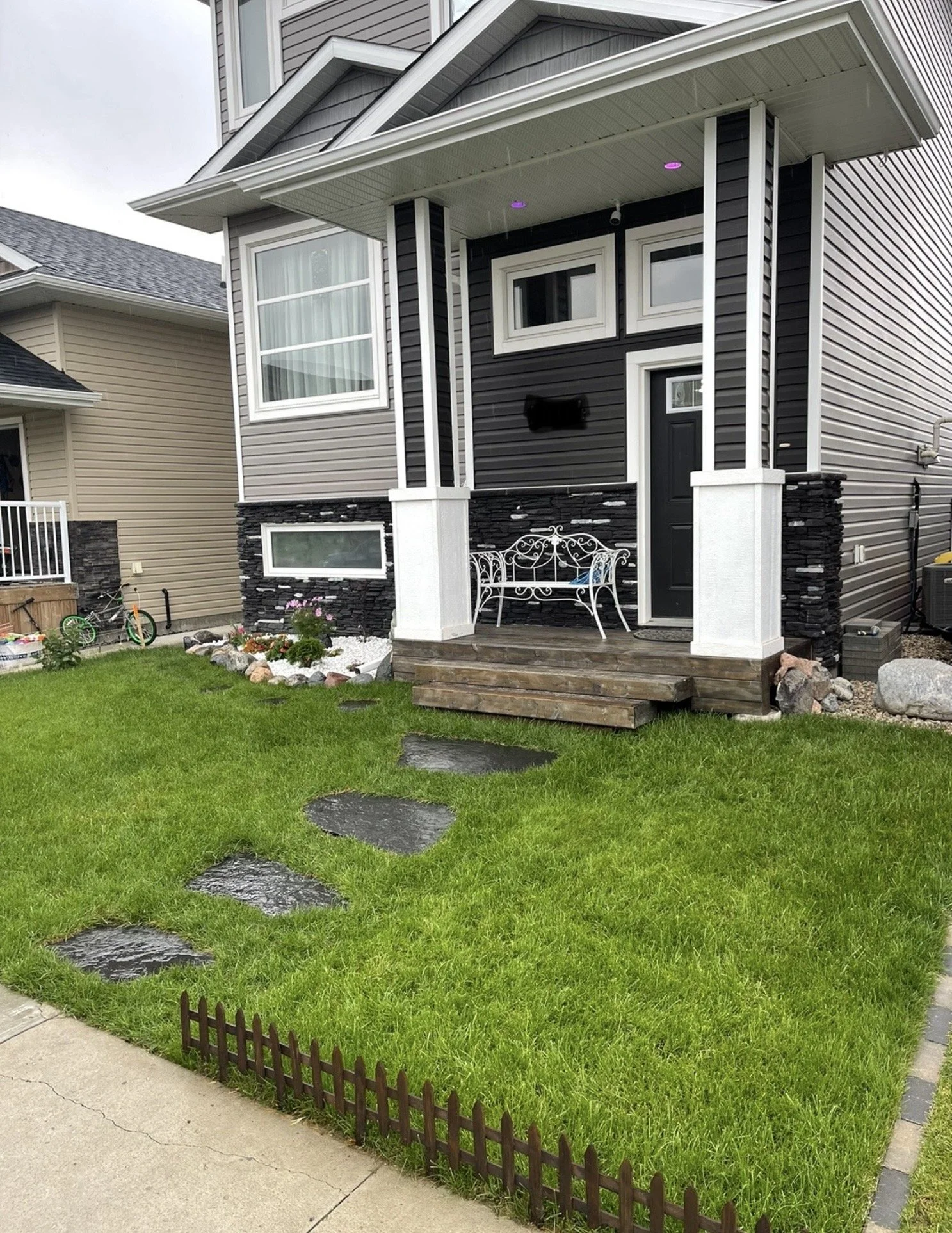 A small front yard with green grass, stepping stones, a tiny brown picket fence, and a porch with steps leading up to a black door on a modern house. The house has gray and black siding, several windows, and a hanging outdoor light.
