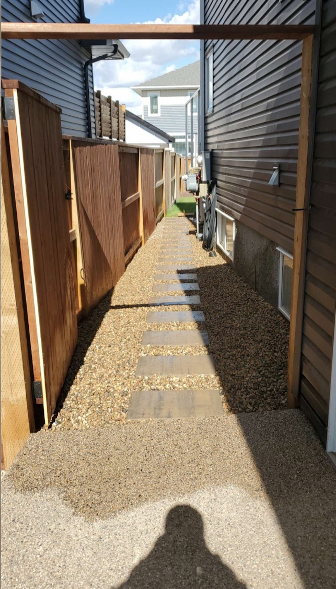A narrow gravel pathway between two houses, with a wooden fence on the left and the house's brown siding on the right. The pathway has concrete stepping stones leading towards the back. There are utility meters attached to the house on the right, and