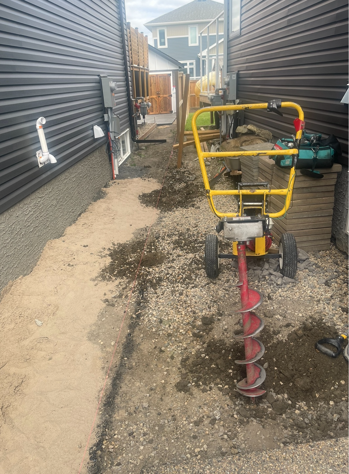 A backyard construction site with an outdoor power auger drill, construction tools, and stacks of paving stones, possibly for a patio or walkway installation. The area is partially excavated with sand and dirt, with a string line marking the work bou