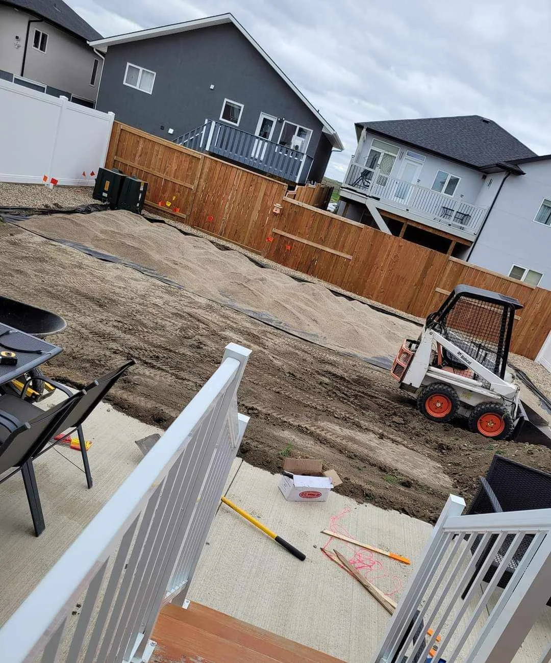Backyard under construction with dirt and a small skid-steer loader, wooden fences, and neighboring houses, with construction tools and supplies on the patio.