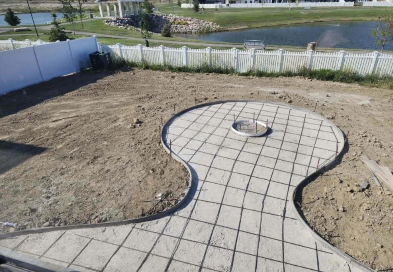 Construction site with a newly poured concrete pathway shaped like a small dog, surrounded by dirt, in a park with a pond, grass, trees, and a white fence in the background.