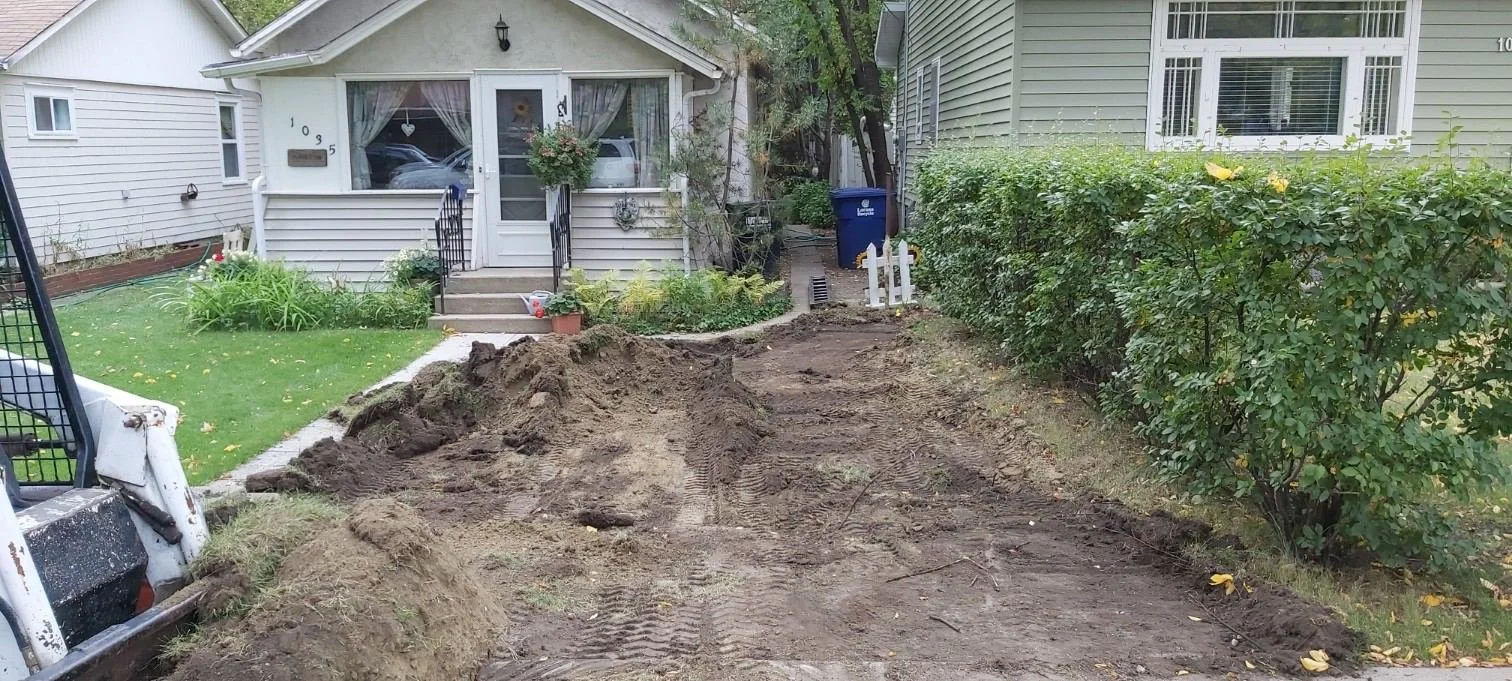 A front yard undergoing driveway renovation, showing dirt and tire marks from construction equipment, with a house and lush shrubbery on either side.