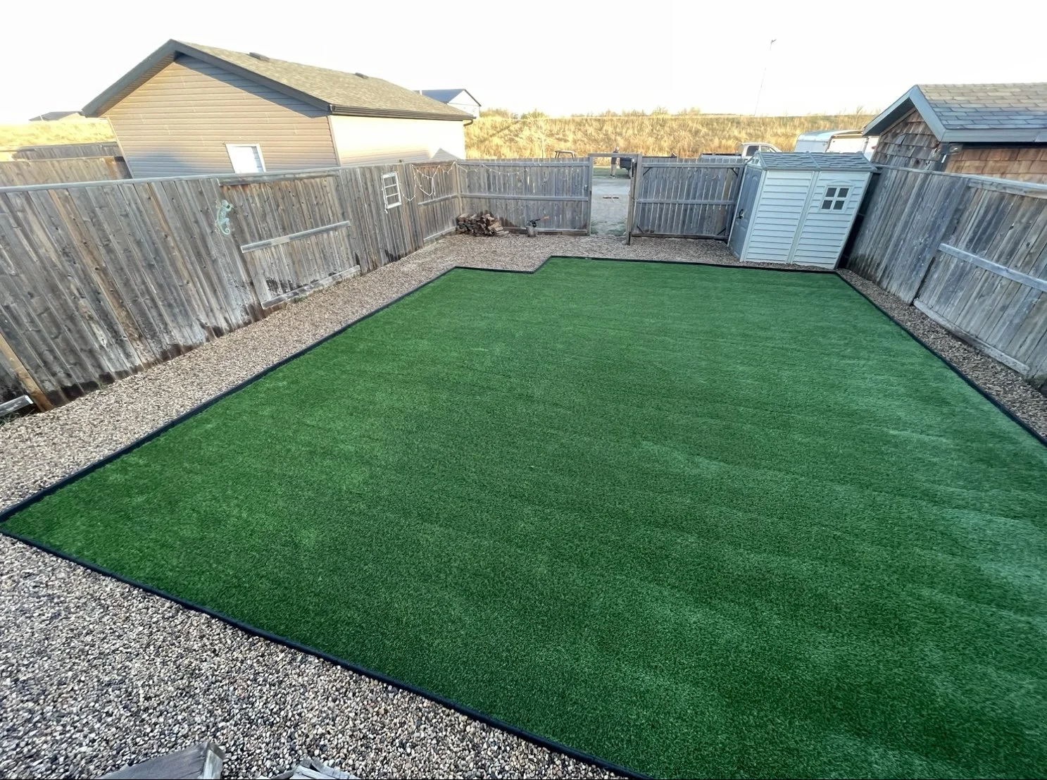 Backyard with a rectangular artificial grass lawn, surrounded by gravel and enclosed by a wooden fence. There is a small storage shed in the corner.