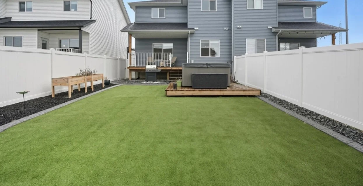 Residential backyard with a green lawn, white privacy fence, wooden deck, and a multi-story blue house in the background.
