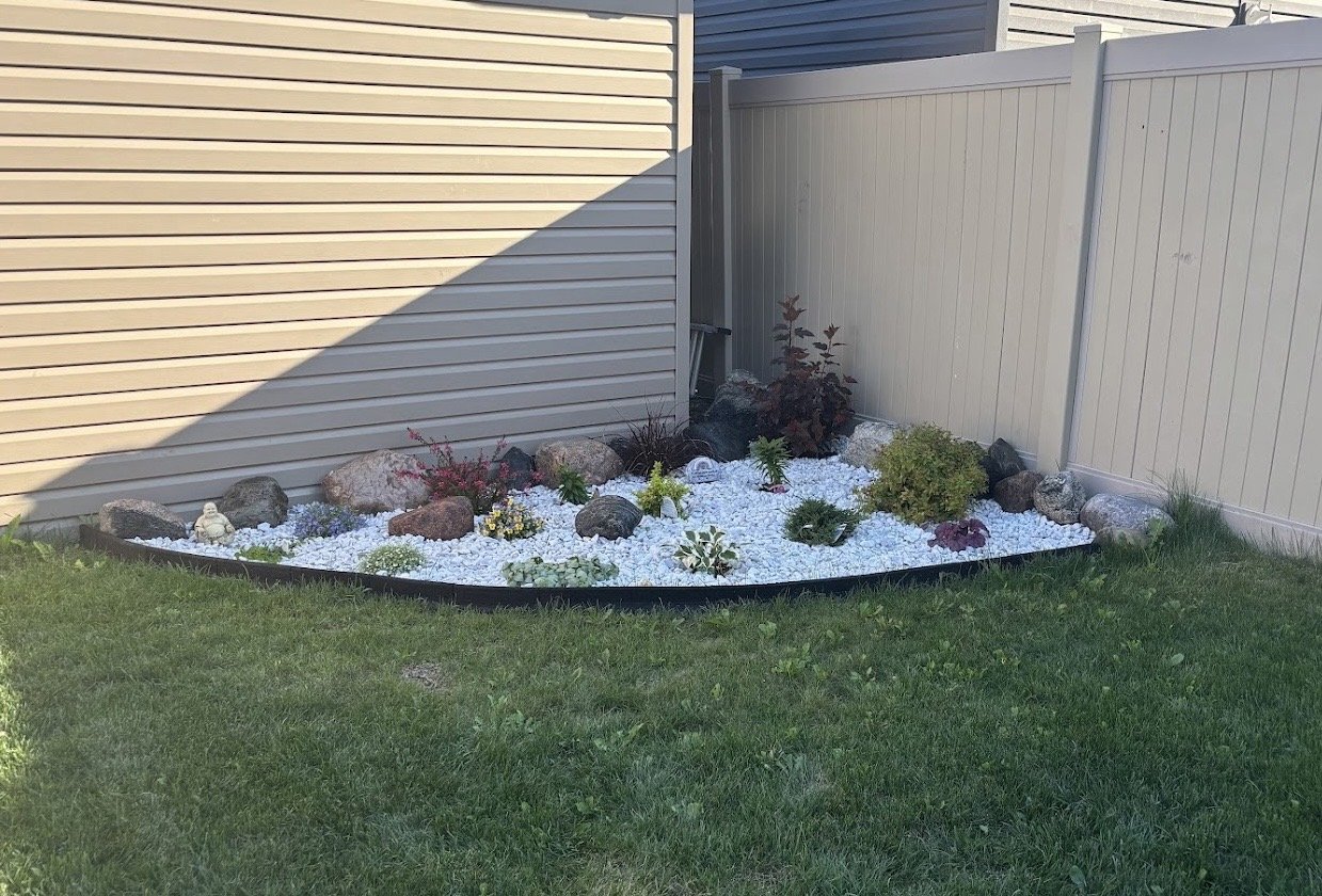 A small garden bed with white gravel, various plants, and rocks, located next to beige and gray fences, with grass in the foreground.