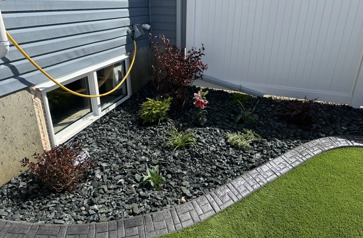 A landscaped garden with black gravel, small plants, and a curved brick border next to a house with gray siding and a white fence.