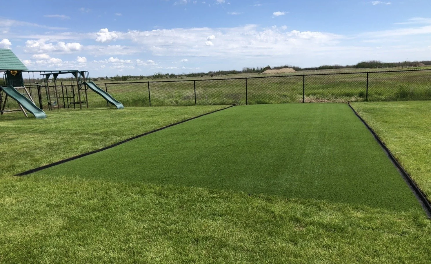 A backyard with a sandbox filled with artificial grass, surrounded by a chain-link fence, in an open field with a playground on the left and a blue sky with scattered clouds.