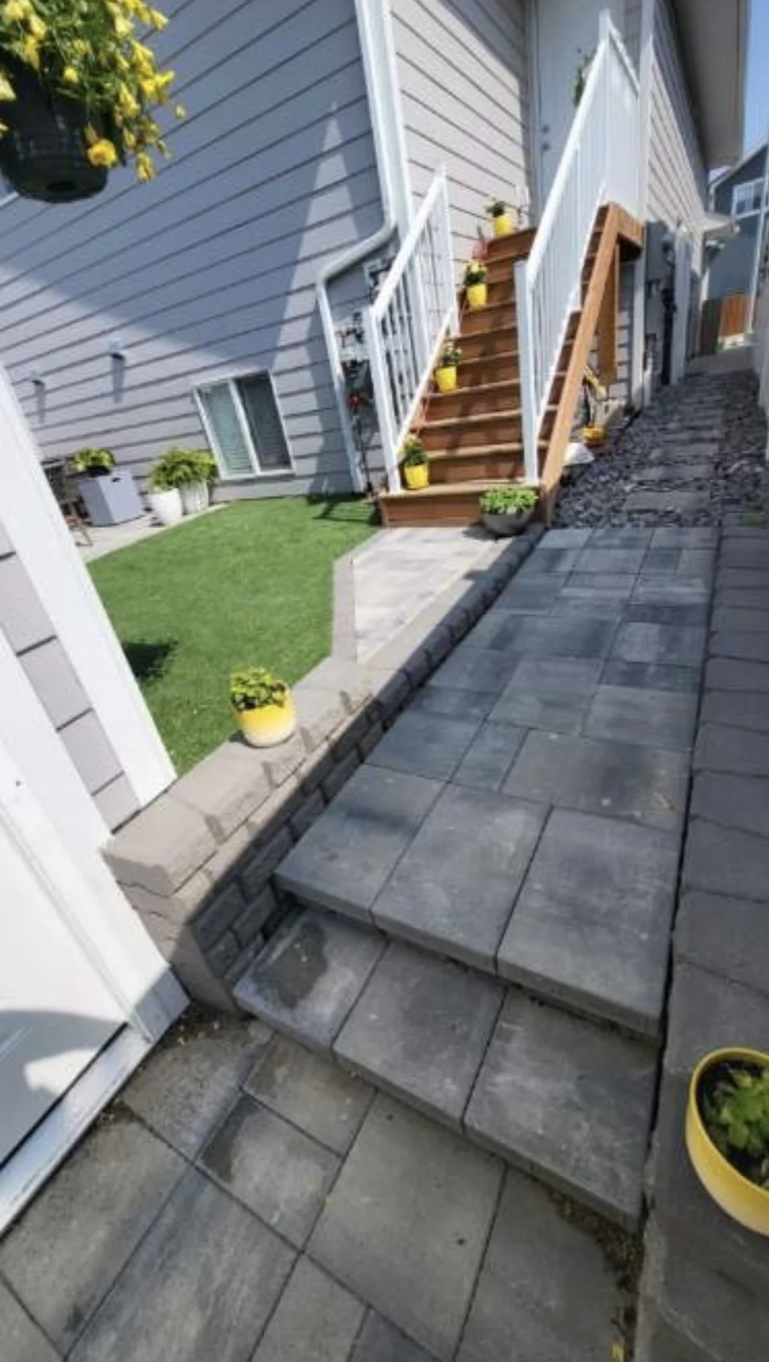 Backyard patio with gray stone steps leading up to a wooden staircase with white railings, surrounding flower pots with yellow and green plants, a small lawn area, and gray siding of a house.