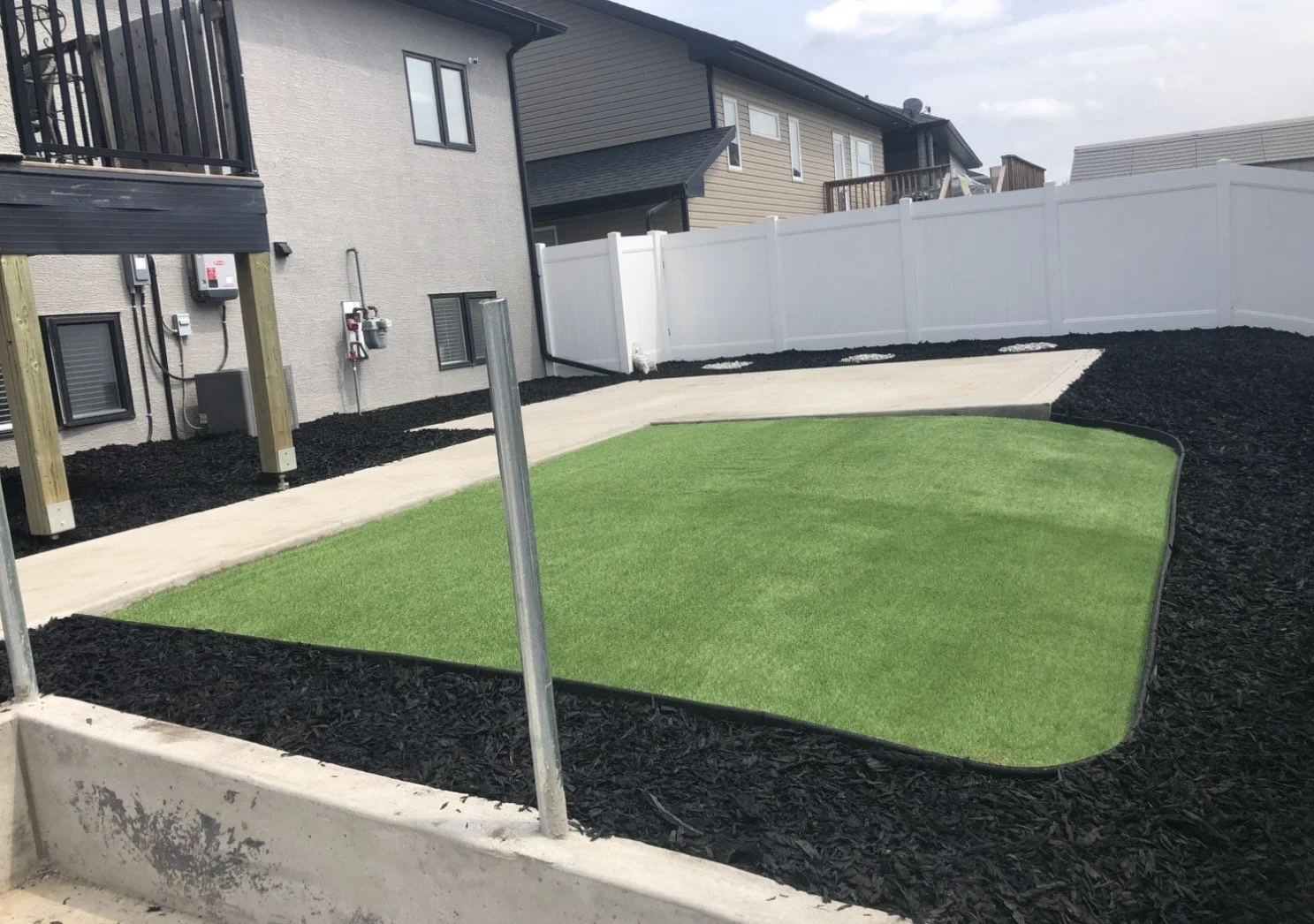 Backyard with artificial grass, concrete pathway, black mulch, and a white privacy fence, with houses in the background.