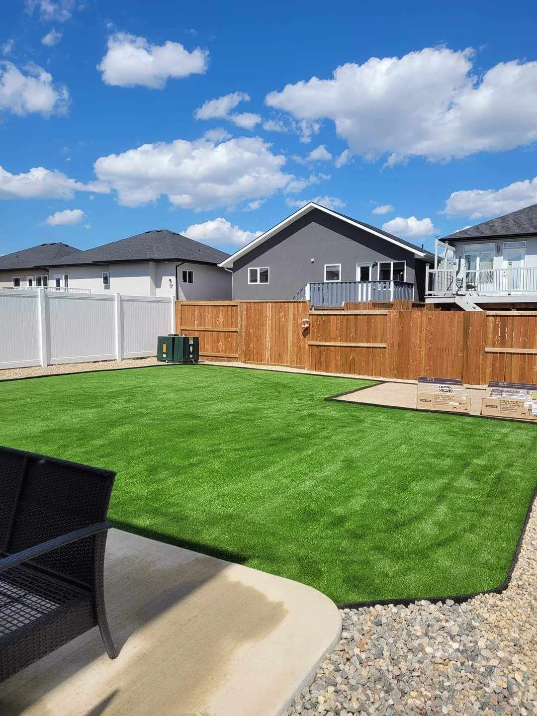Backyard with artificial grass, wooden and white fences, patio furniture, and neighboring houses under a blue sky with clouds.