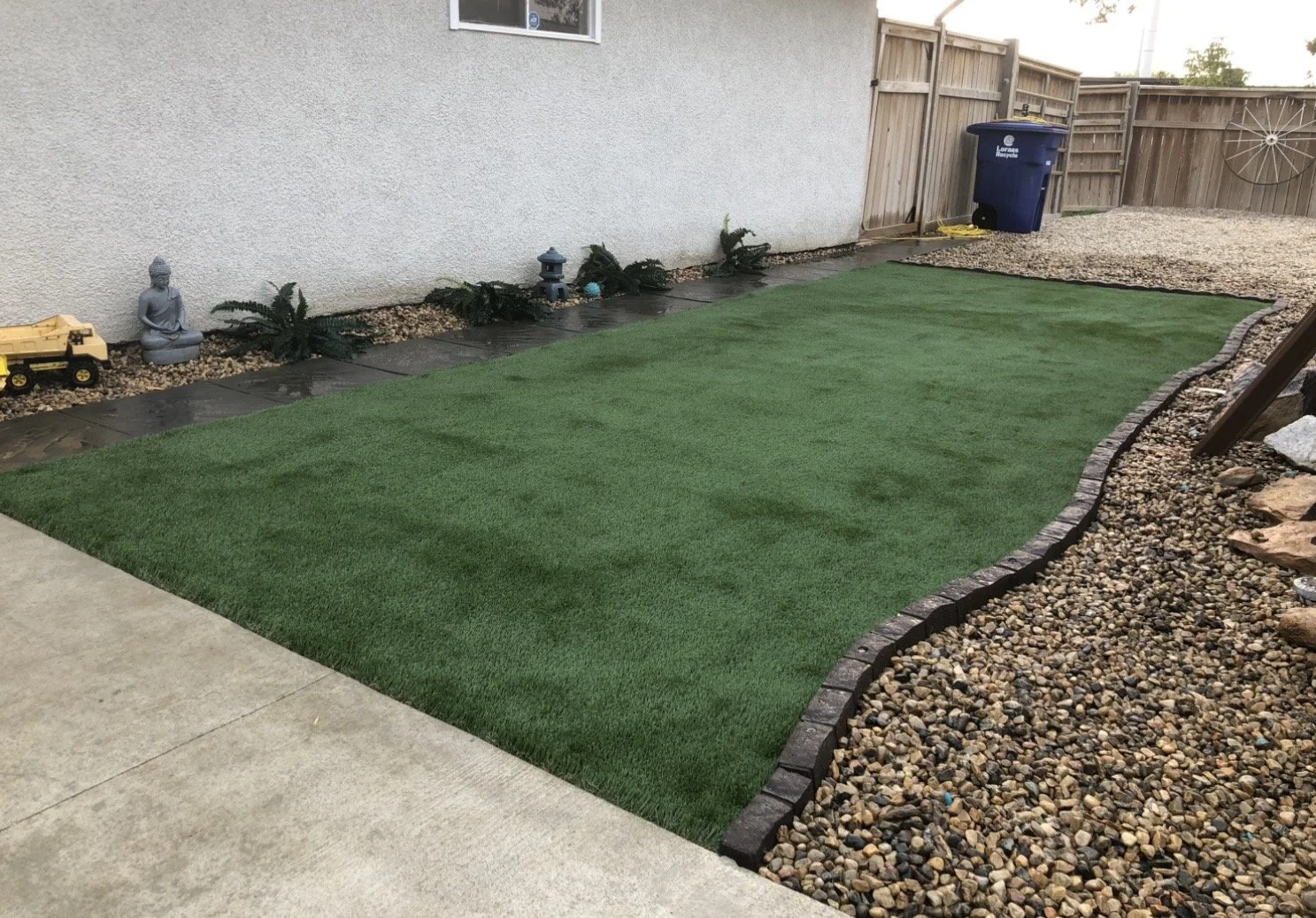 Backyard with artificial grass, surrounding rocks, and small plants along the house wall. Fenced area with a trash bin and decorative wheel in the corner.