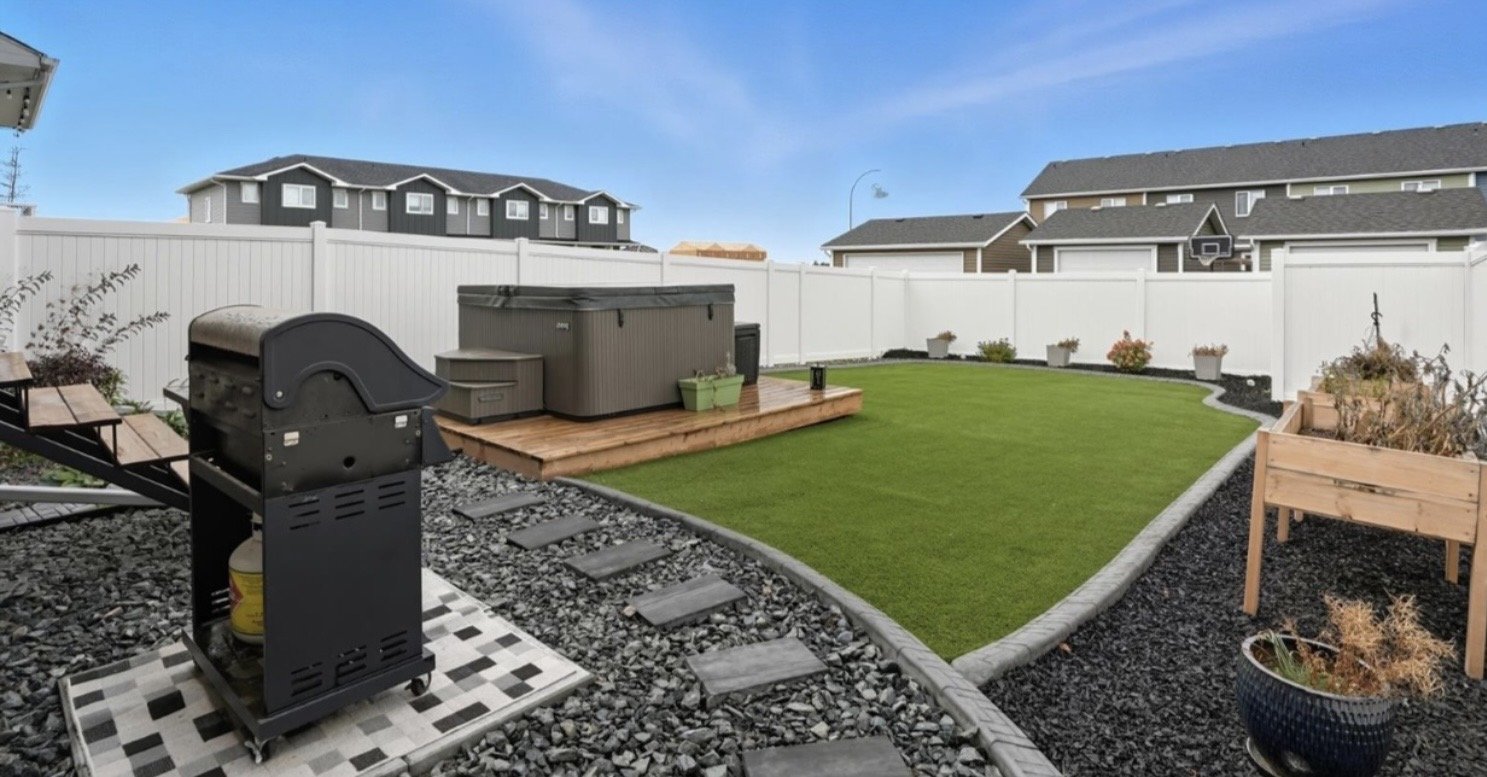 Backyard with a green lawn, a hot tub on a wooden deck, a barbecue grill on gravel, a row of potted plants against a white fence, and neighboring houses in the background.