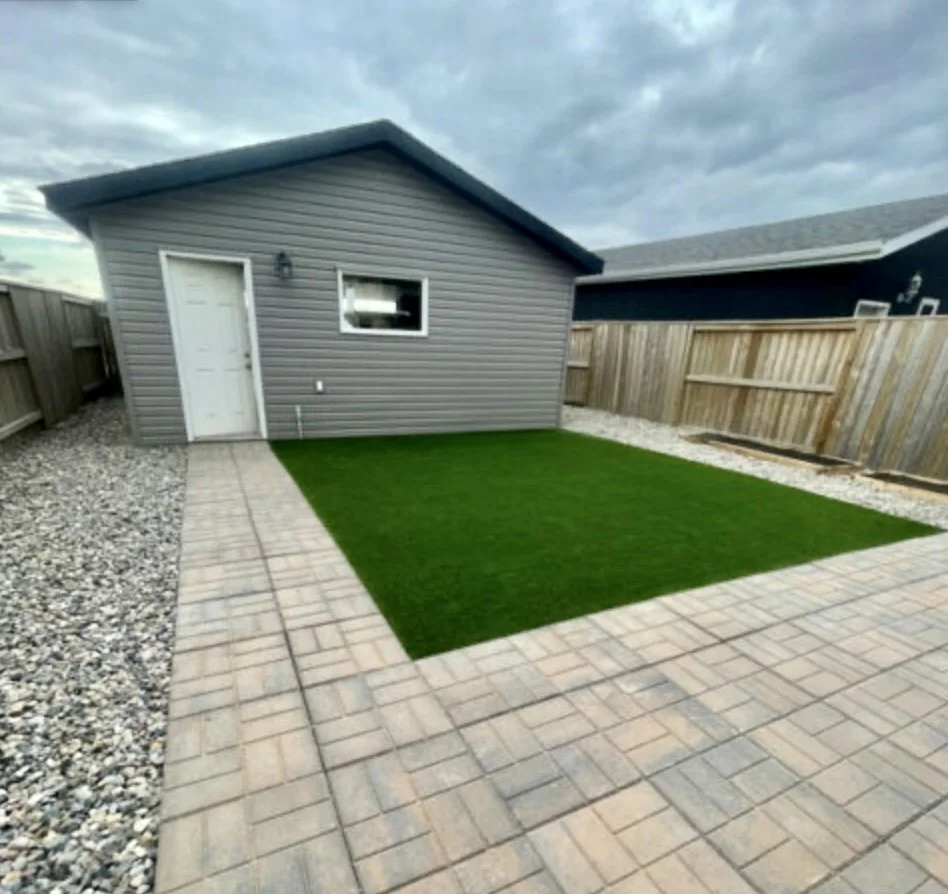Backyard with a small house, a patch of artificial grass, a paved area, a gravel border, and a wooden fence.