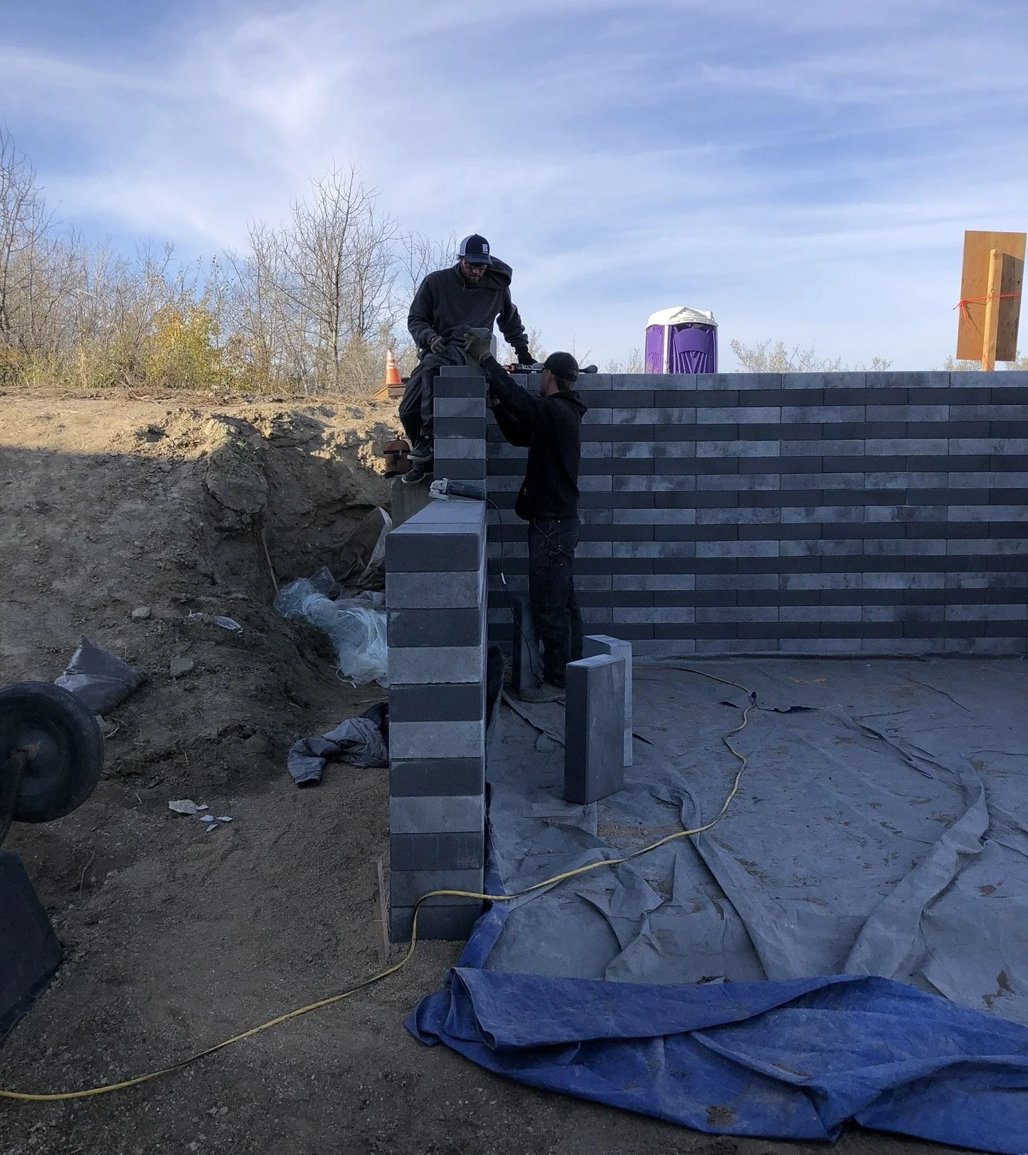 Two men working on constructing a brick wall outdoors under a clear sky. One man is standing on the ground, helping the other who is on the wall. There are construction materials and equipment around, including a purple portable toilet in the backgro