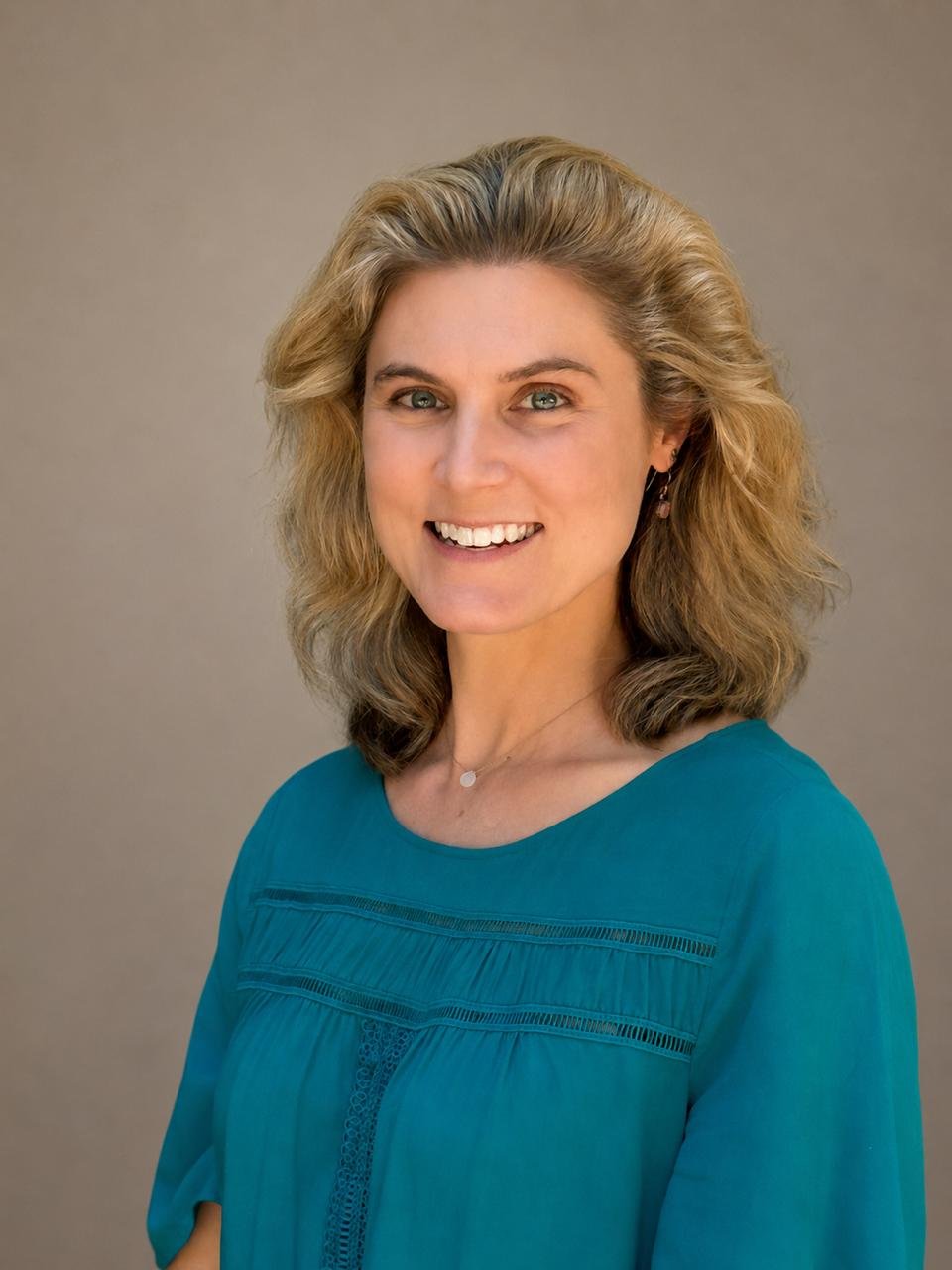 A smiling woman with shoulder-length blonde hair, wearing a teal top and earrings, posing against a neutral background.