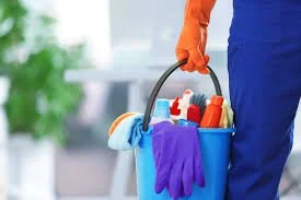 Person holding a blue cleaning bucket with orange gloves and various cleaning supplies inside.