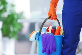 Person in blue pants and orange gloves holding a cleaning bucket filled with cleaning supplies like spray bottles and cloths.