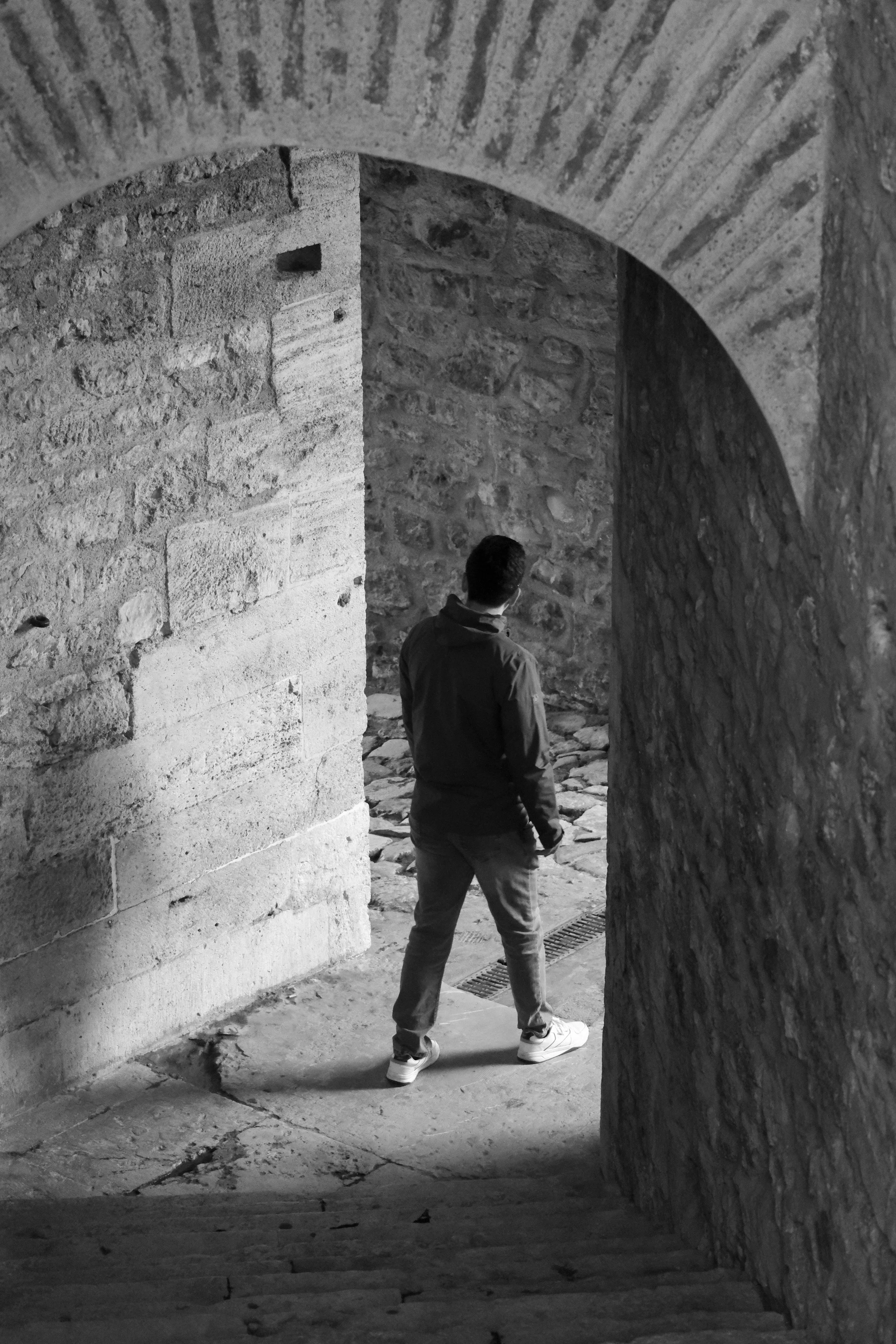 Homme vu de dos dans une ruelle pavée en pierre, entouré de murs en pierre et en brique, vue en noir et blanc