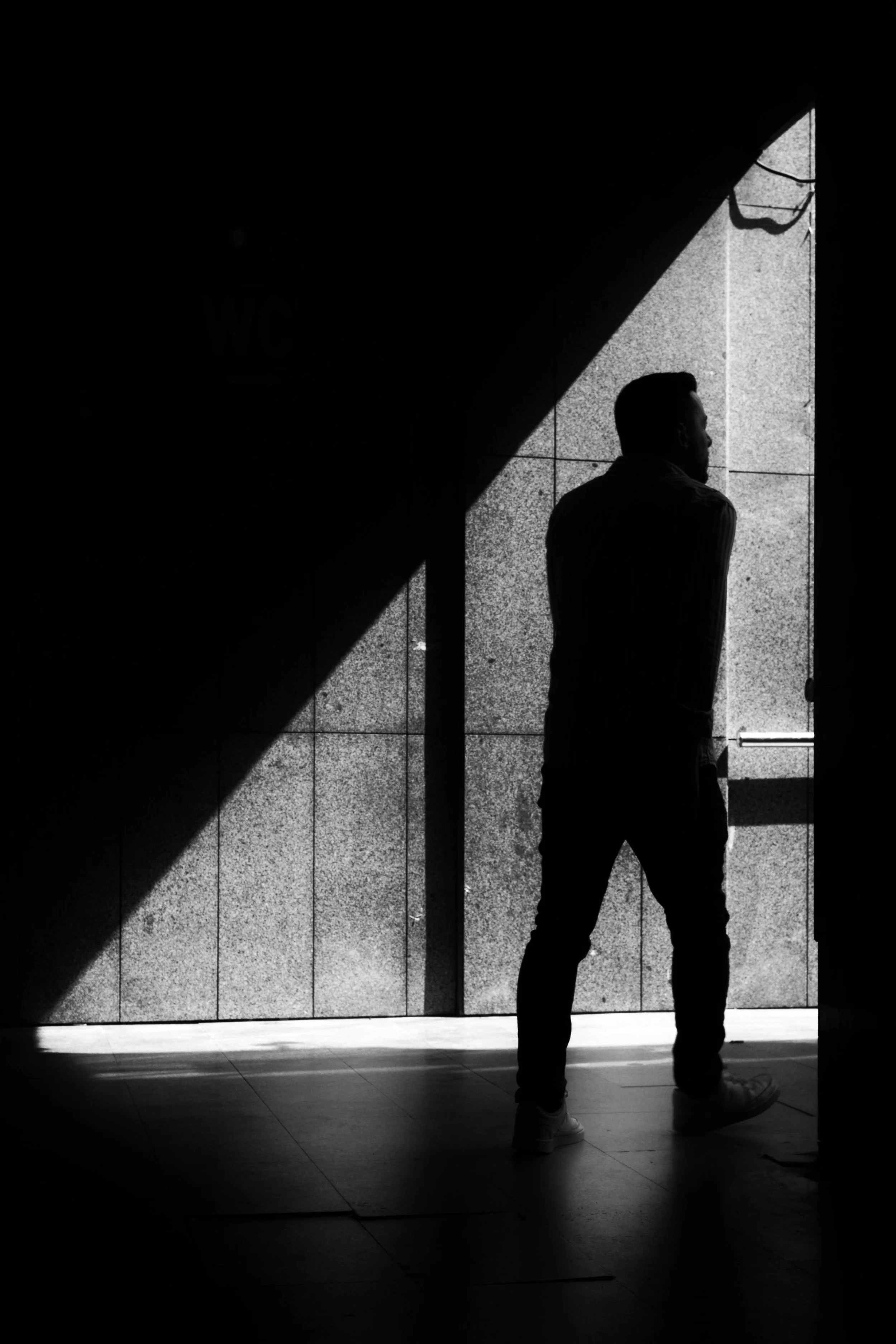 homme debout dans une zone partiellement éclairée, avec un mur en arrière-plan, en noir et blanc