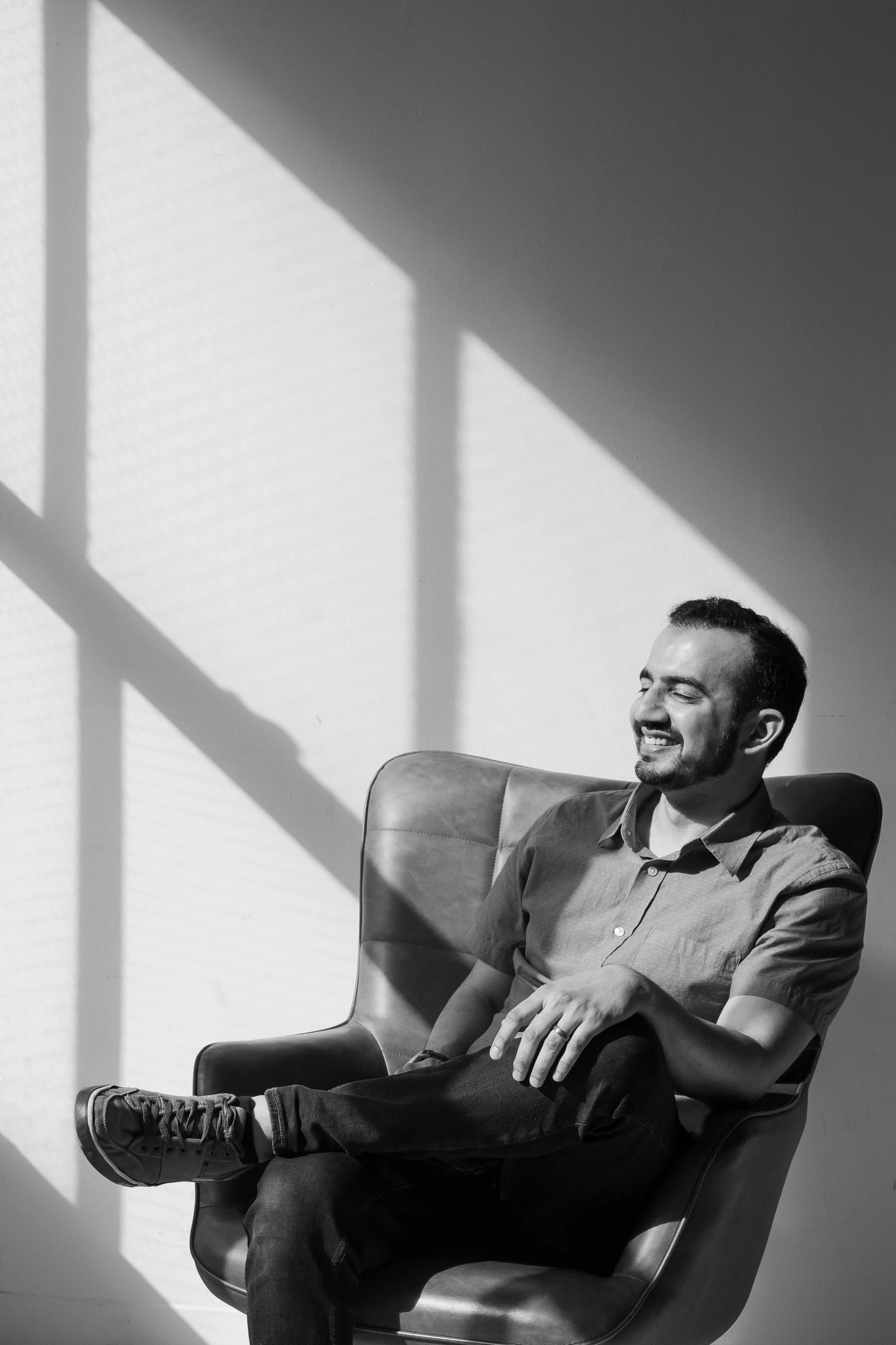 Un homme assis dans une chaise en cuir, souriant et semblant détendu, avec des ombres de fenêtres projetées sur le mur derrière lui