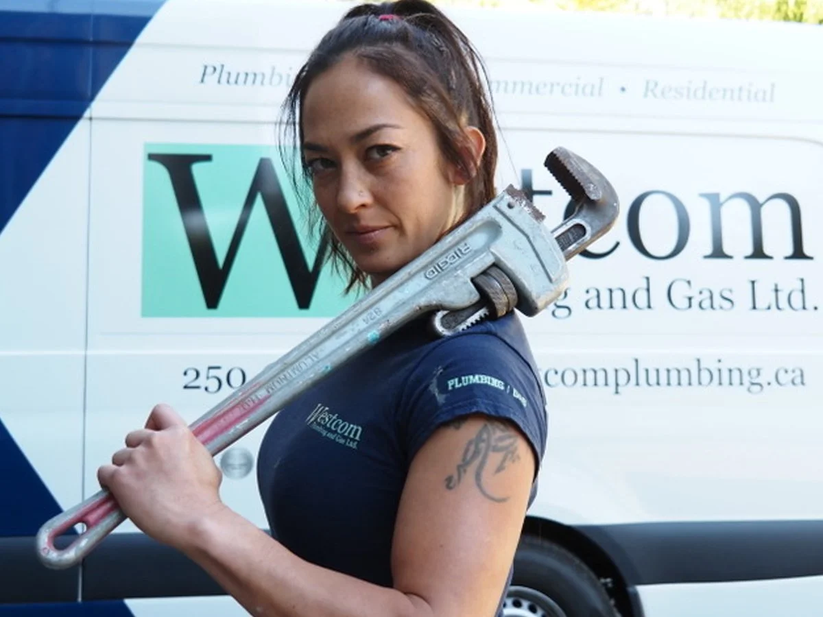 A woman holding a large pipe wrench over her shoulder, standing in front of a plumbing company van.