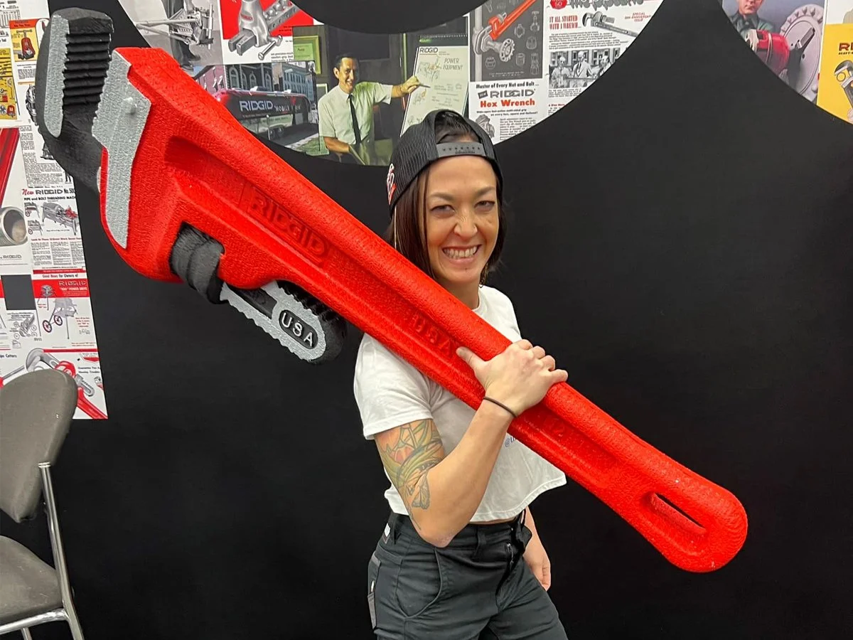 Woman holding a giant red foam wrench, smiling, wearing a backward black cap, with tattoos on her left arm, in front of a black background with posters and advertisements for tools.