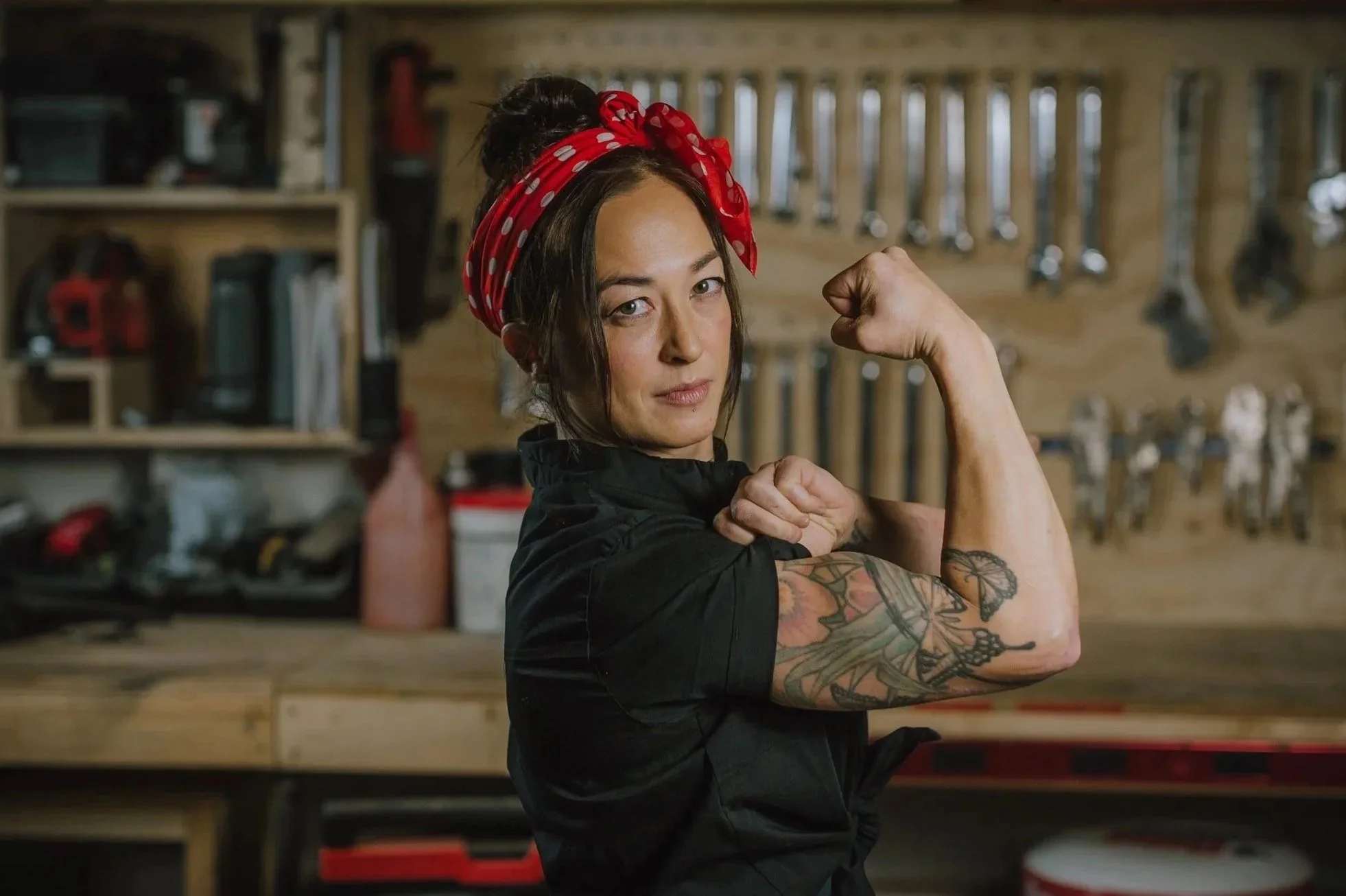 A woman with dark hair in a bun, wearing a red polka-dot headscarf and black clothing, flexing her right arm to show a tattooed bicep in a workshop with tools hanging on a wooden wall in the background.