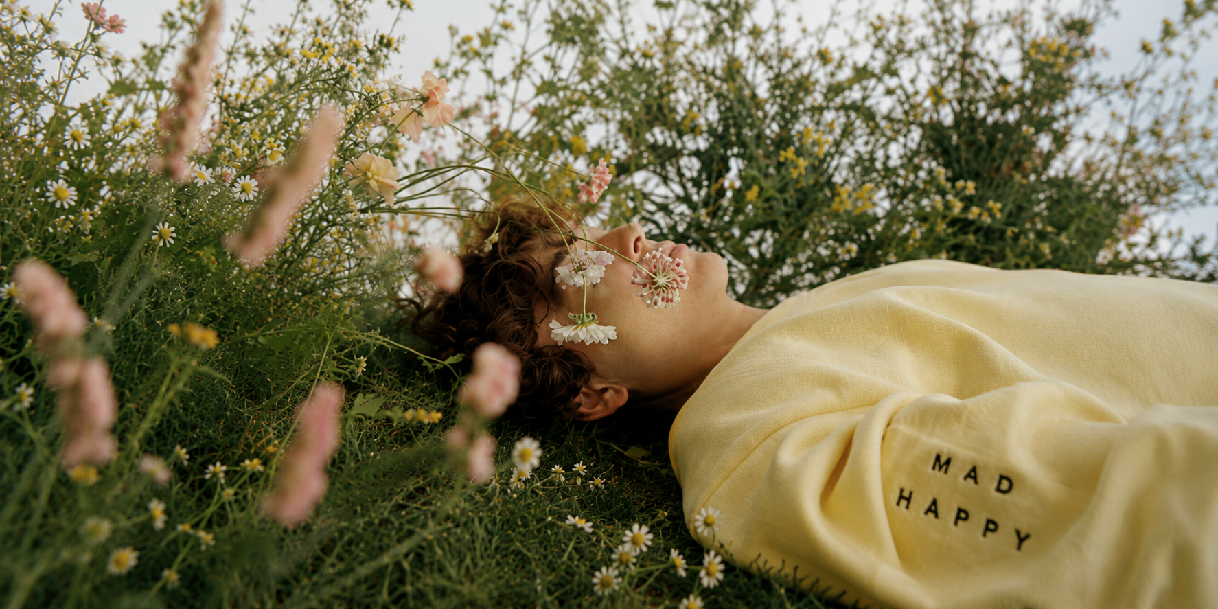 A woman lying on the ground in a field of flowers, with her face turned sideways, wearing a yellow shirt with the words "Mad Happy" on it, surrounded by blooming flowers and greenery.