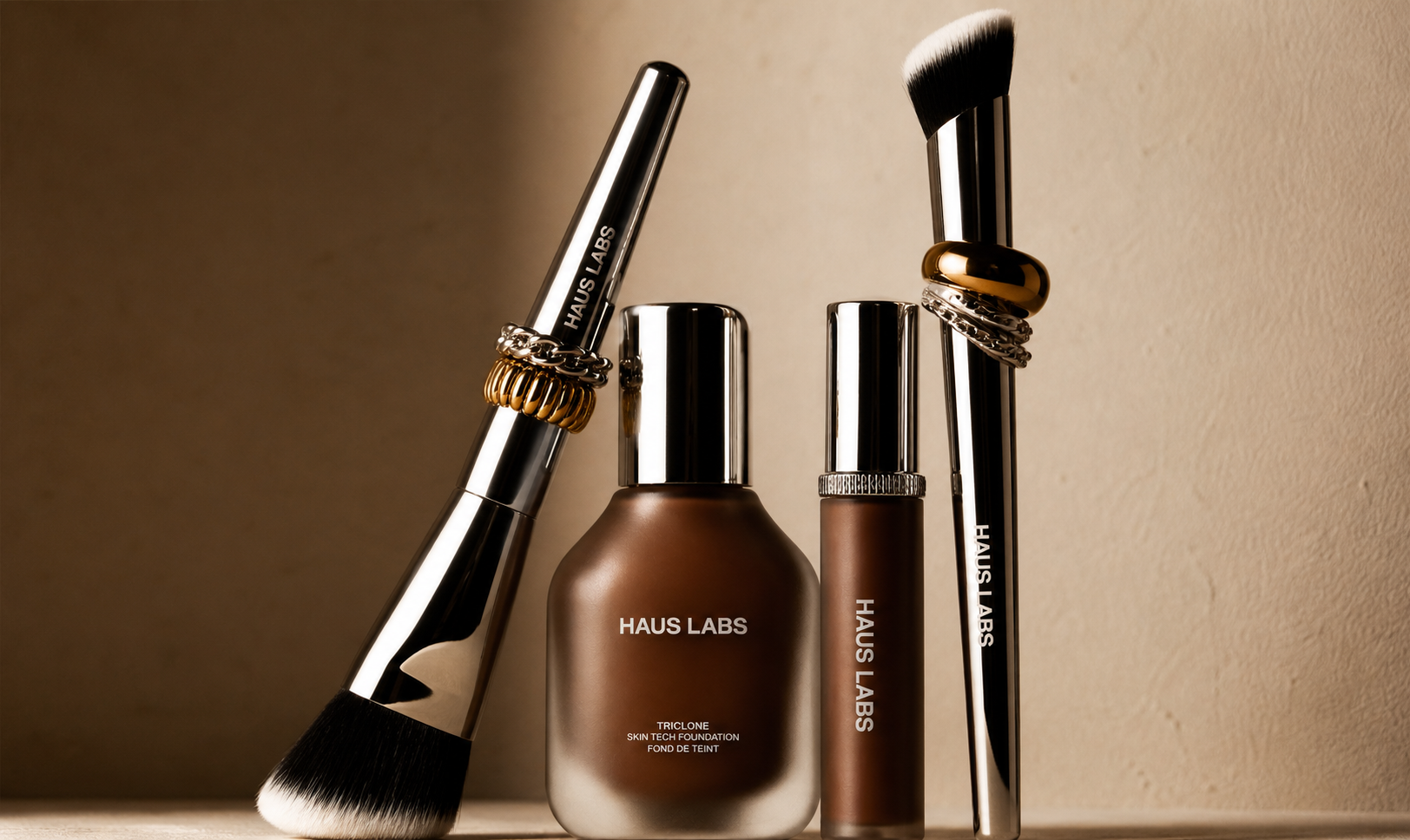 Four makeup brushes and two bottles of foundation makeup products from Haus Labs, displayed against a beige background.