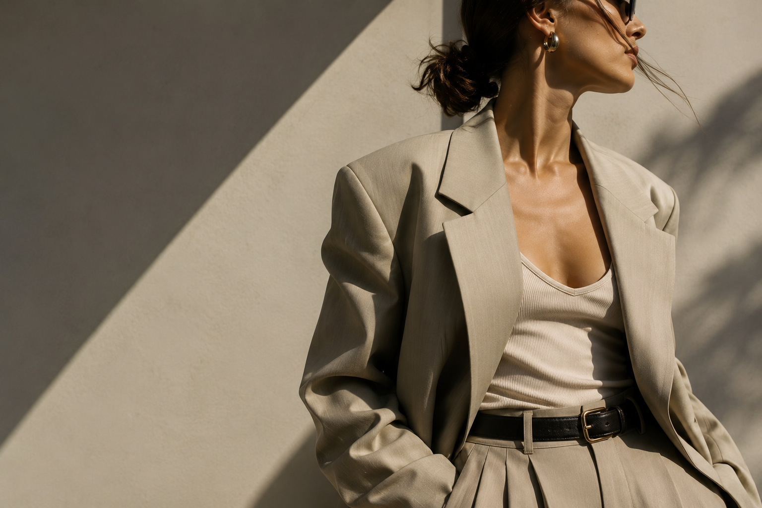 Woman in beige blazer and pleated pants with a belt, wearing hoop earrings, standing against a light-colored wall, looking to the side.