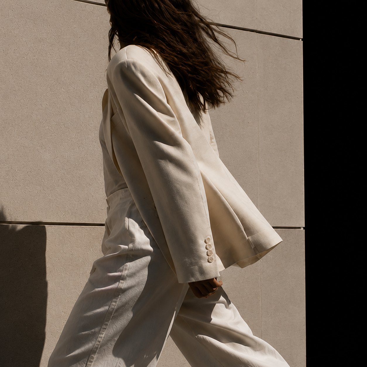 Person with wavy hair wearing a cream-colored suit walking outdoors against a light stone wall with a black section.