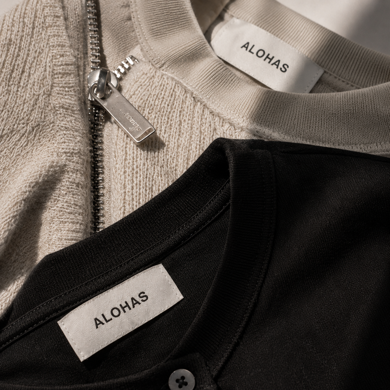 Close-up of two clothing items, one in beige with a silver zipper, and the other in black, both with white labels reading 'ALOHA S'.