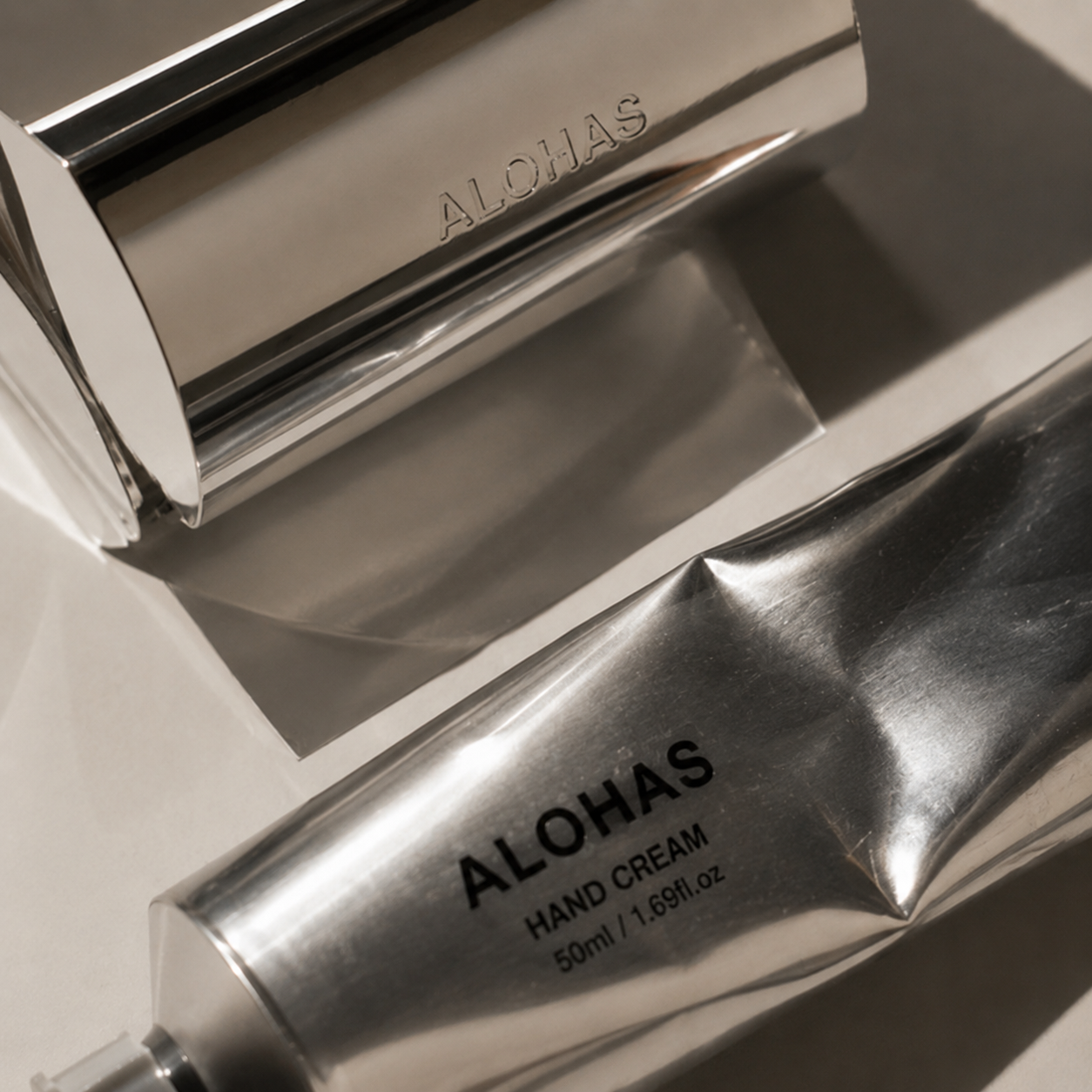 A stainless steel tube labeled 'ALOHA HAND CREAM 50ml' and a matching stainless steel tube labeled 'ALOHA', placed on a flat surface.